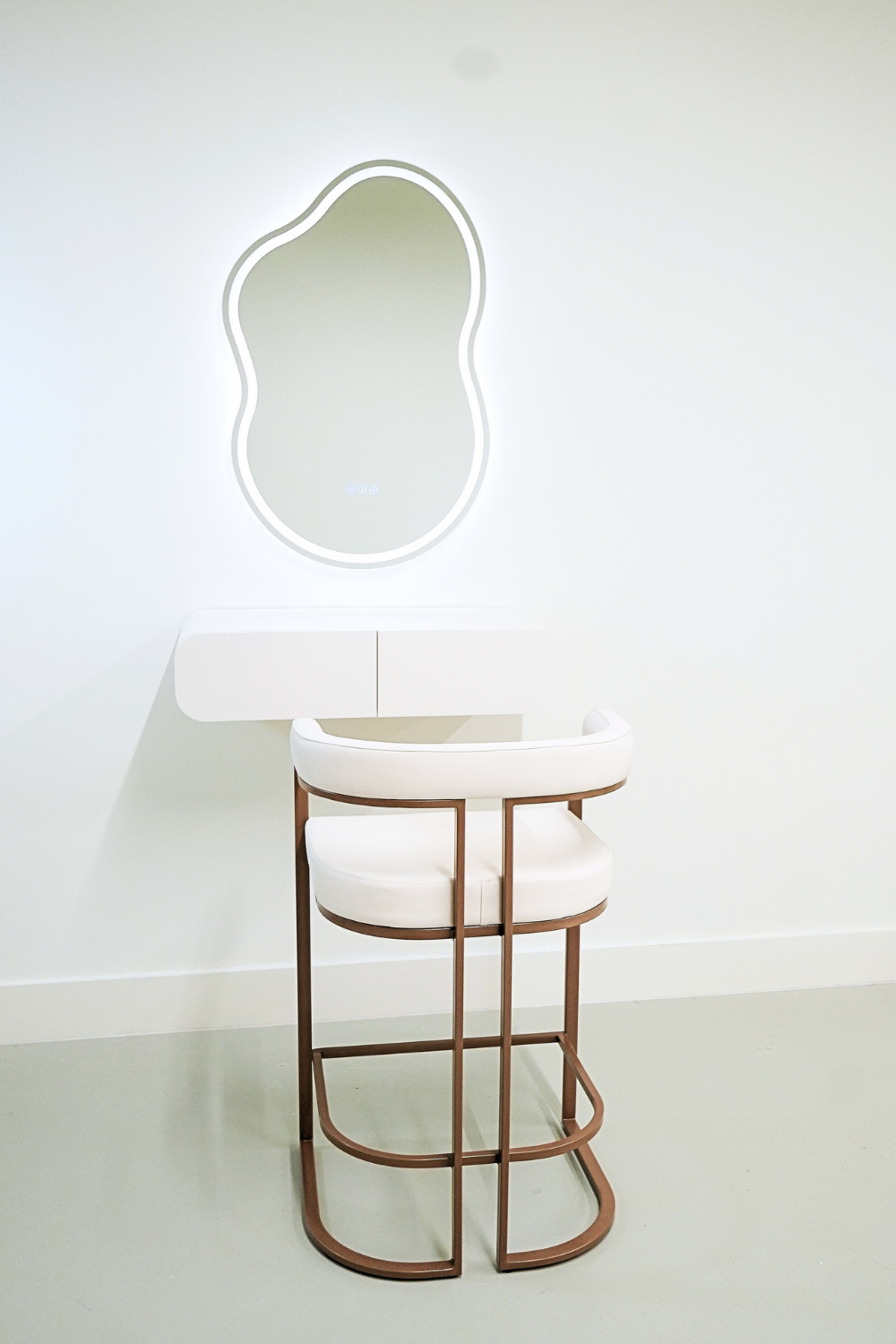 A modern vanity setup features a uniquely shaped illuminated mirror above a white drawer unit and a minimalist white cushioned chair with a bronze frame.