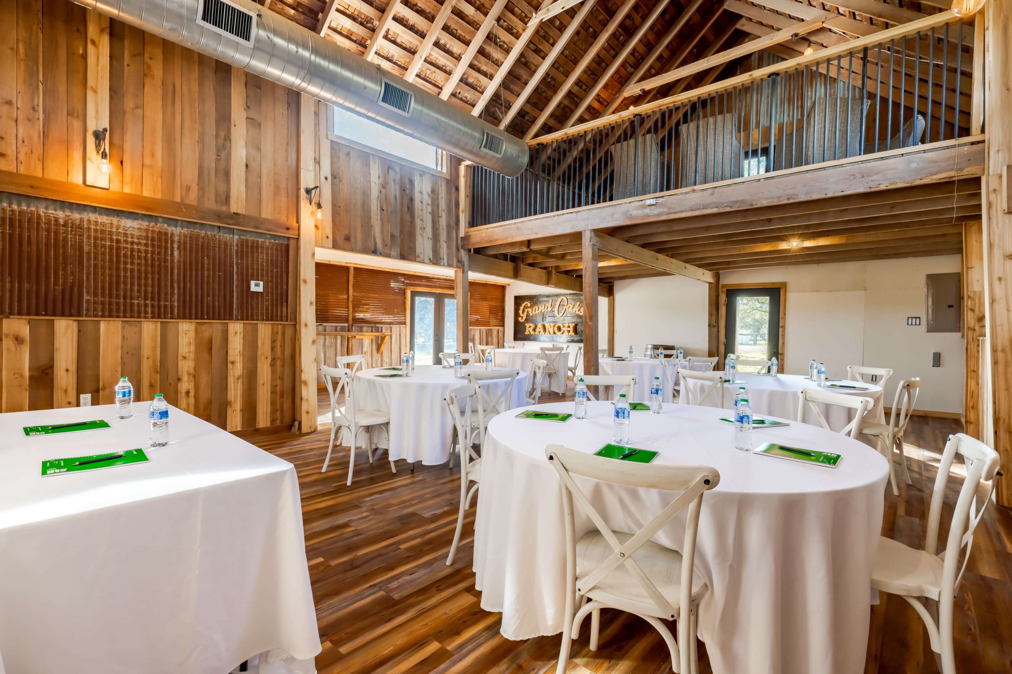 A wooden interior space set up for a meeting or event, featuring round tables with white tablecloths and green placemats.