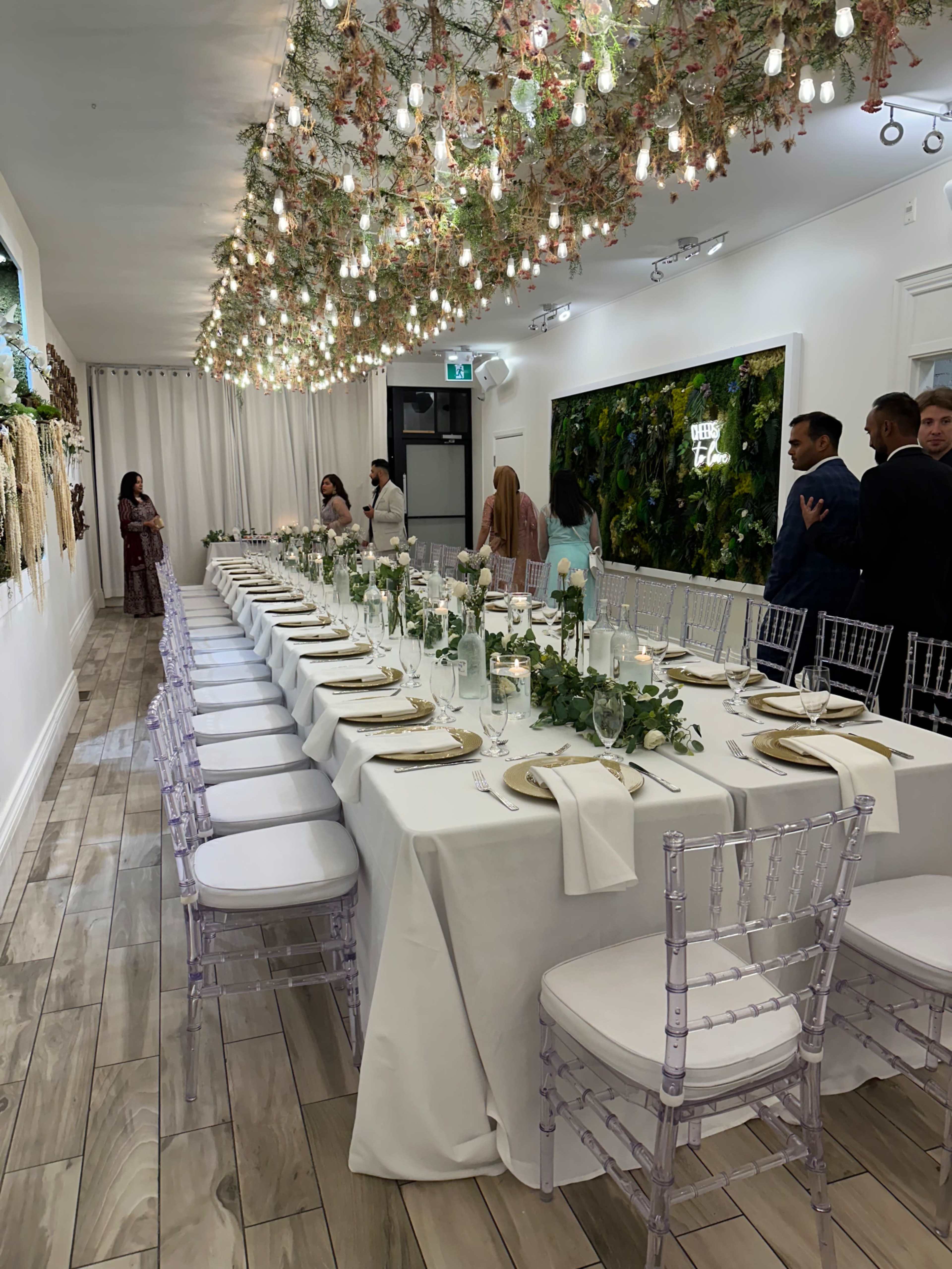 A long, elegantly set dining table with white linens and floral decorations is arranged in a well-lit room adorned with hanging greenery and string lights.