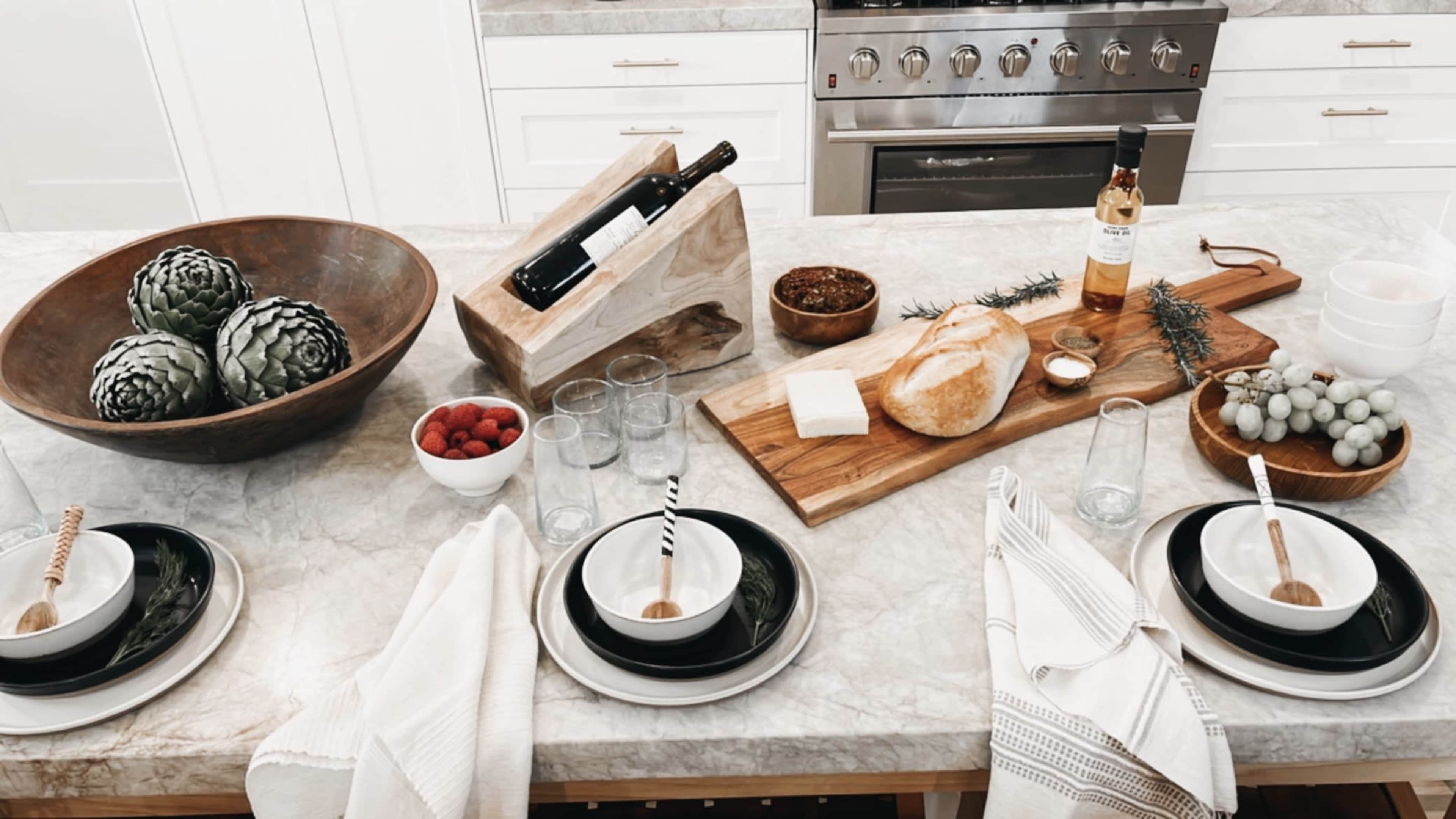 A kitchen table is set with black and white dishware, glasses, and a variety of food items, including bread, raspberries, and a bottle of wine, arranged on a marble countertop.