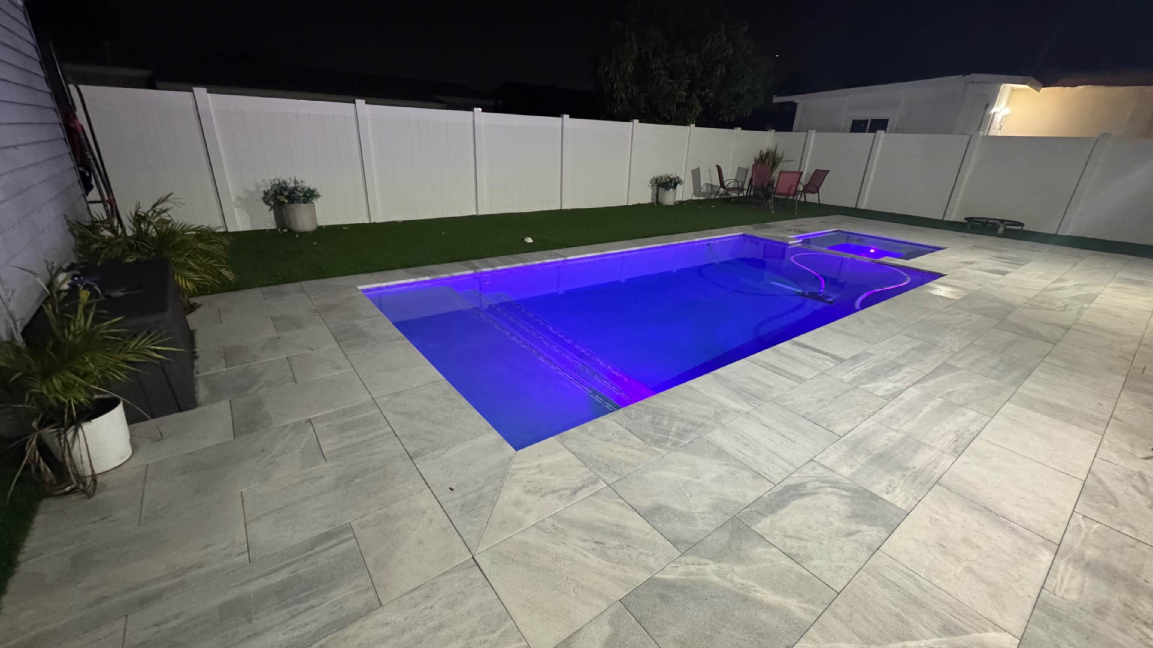 The image shows a rectangular swimming pool with purple lighting, surrounded by a stone patio and a white fence.