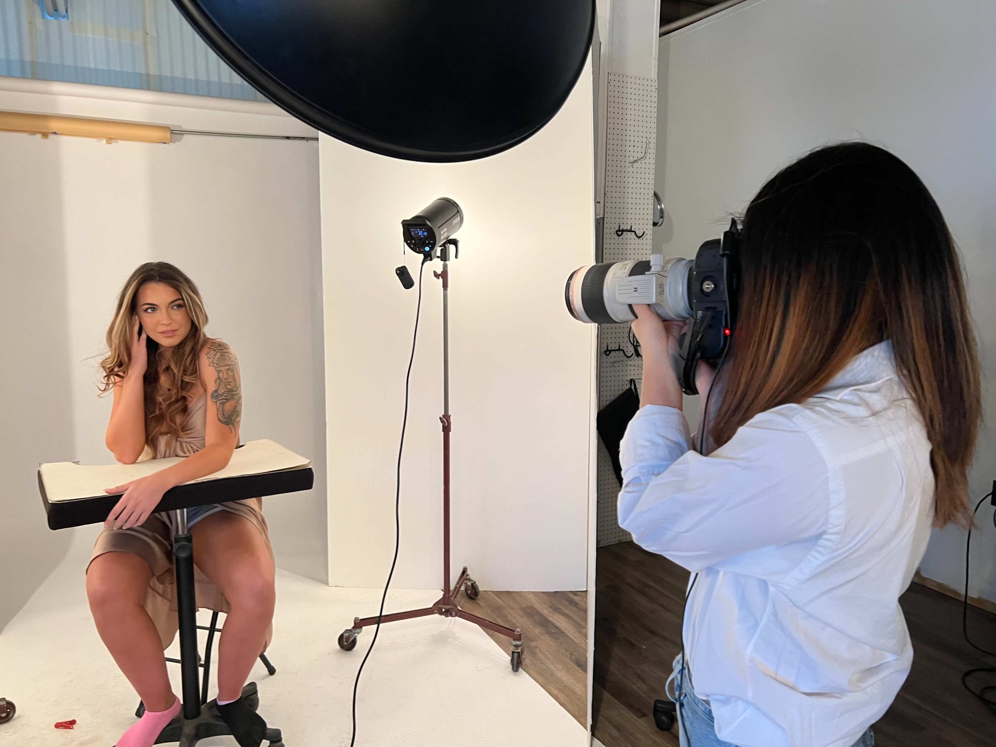 A photographer is capturing an image of a model sitting on a stool in a studio setup.