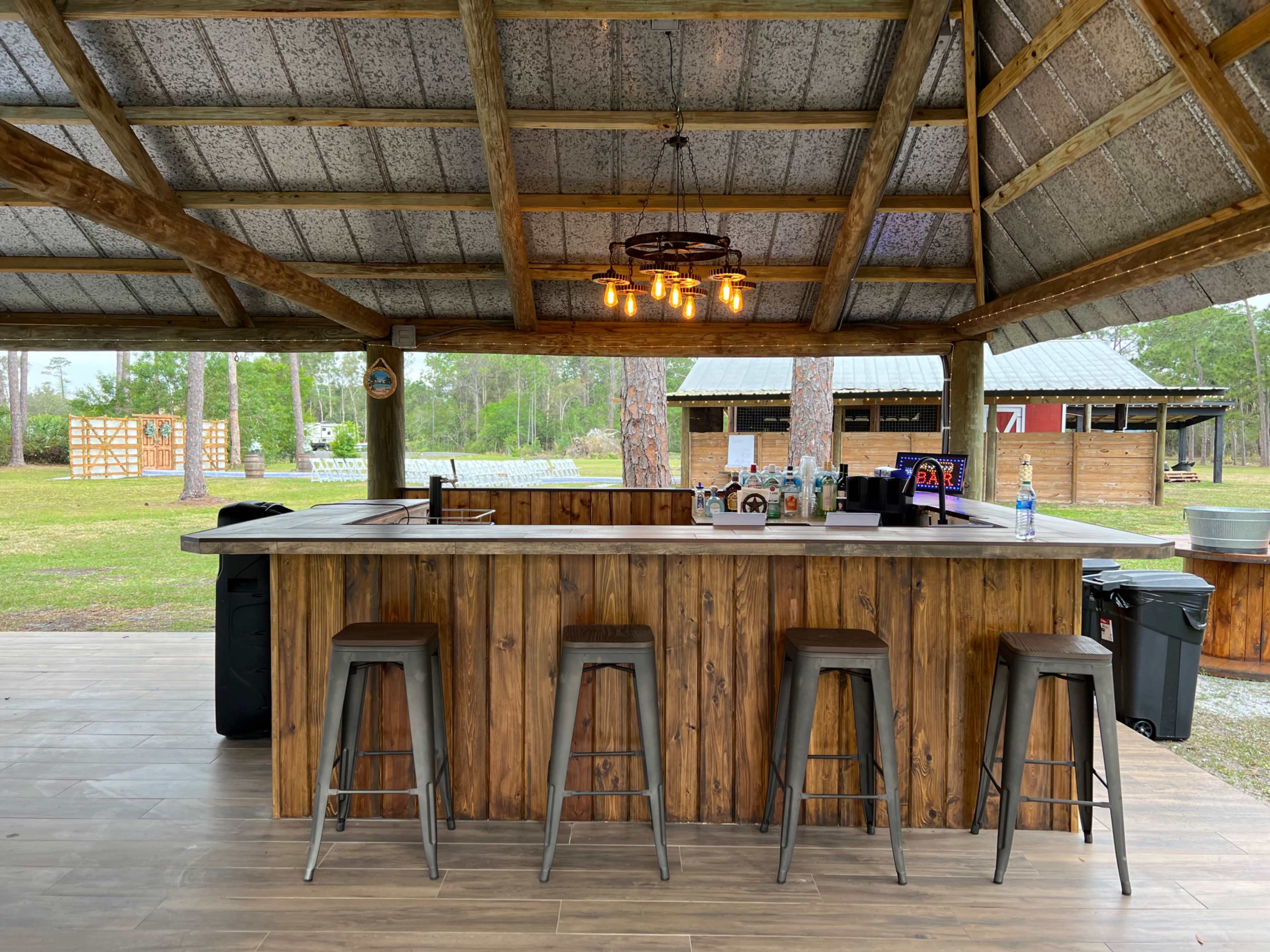 The image shows a rustic outdoor bar with a wooden counter and high stools, situated under a covered shelter surrounded by trees and a grassy area.
