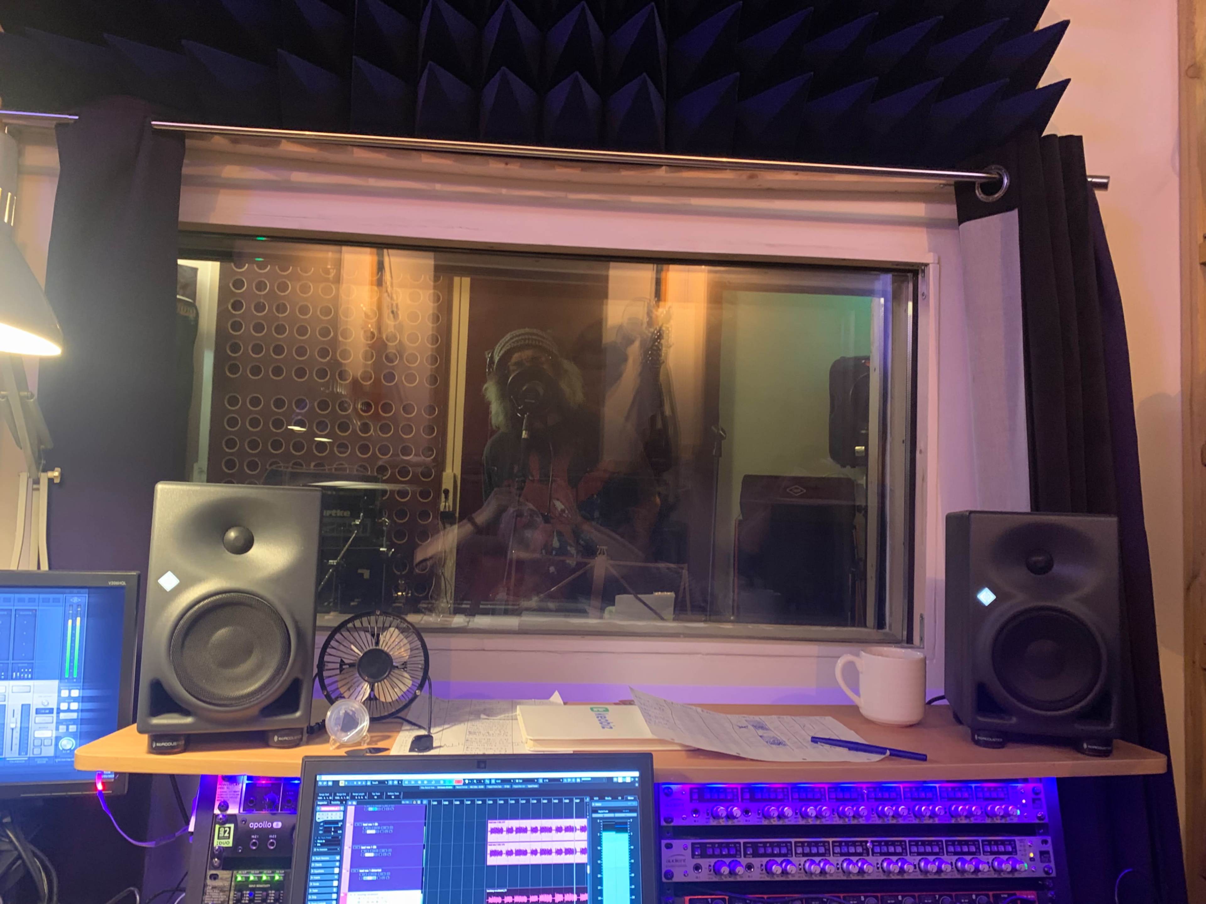 A recording studio with large speakers, a computer workstation, and a window displaying a person playing an instrument inside the sound booth.
