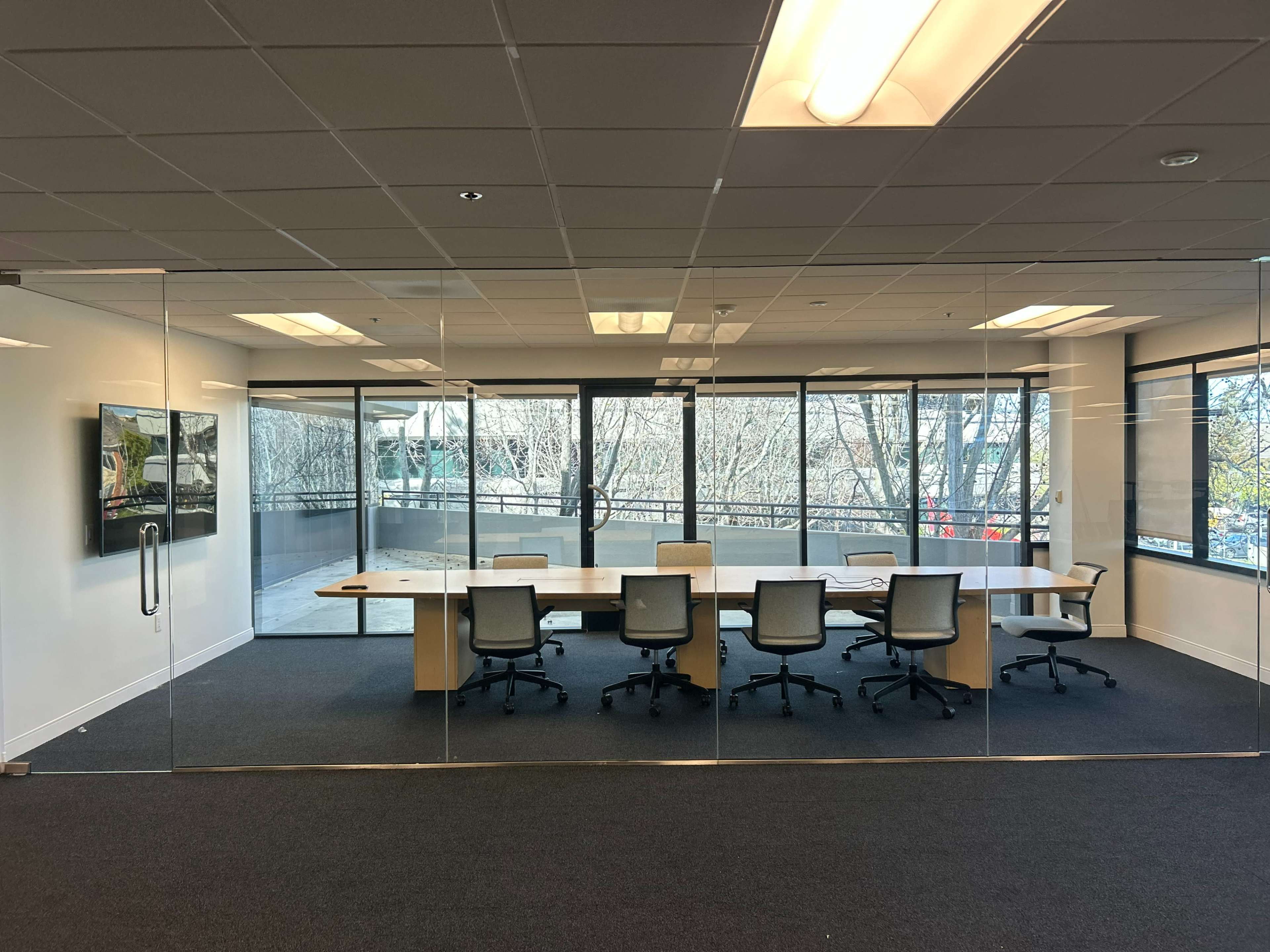The image shows a modern conference room with a large table and several chairs, enclosed by glass walls, overlooking an outdoor view.