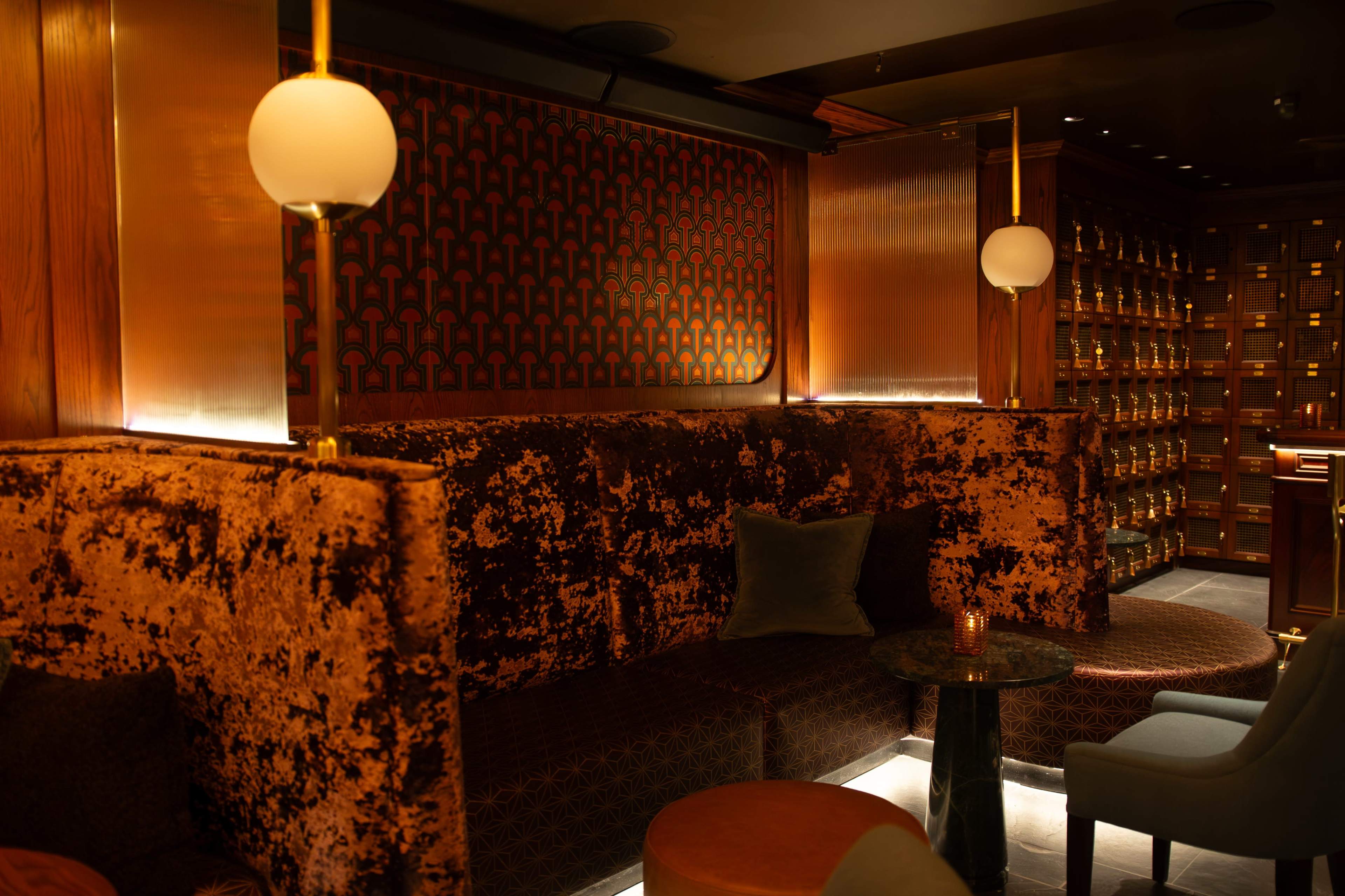 The image shows a stylish lounge area featuring plush velvet seating, warm lighting, and decorative wall patterns.