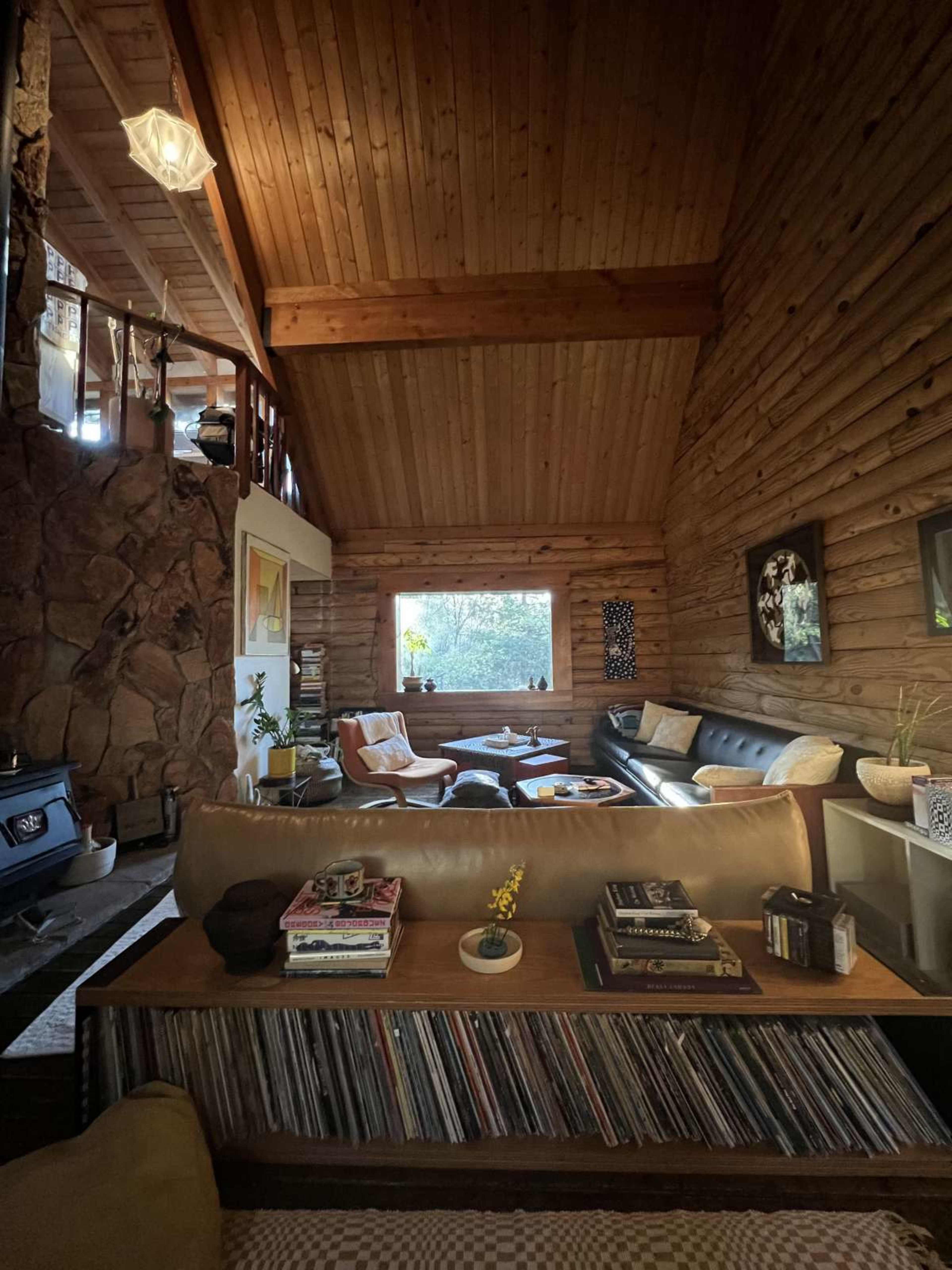 The living room features a high wooden ceiling, a combination of leather and fabric seating, and a large window allowing natural light to pour in, alongside a collection of books and records on a shelf.