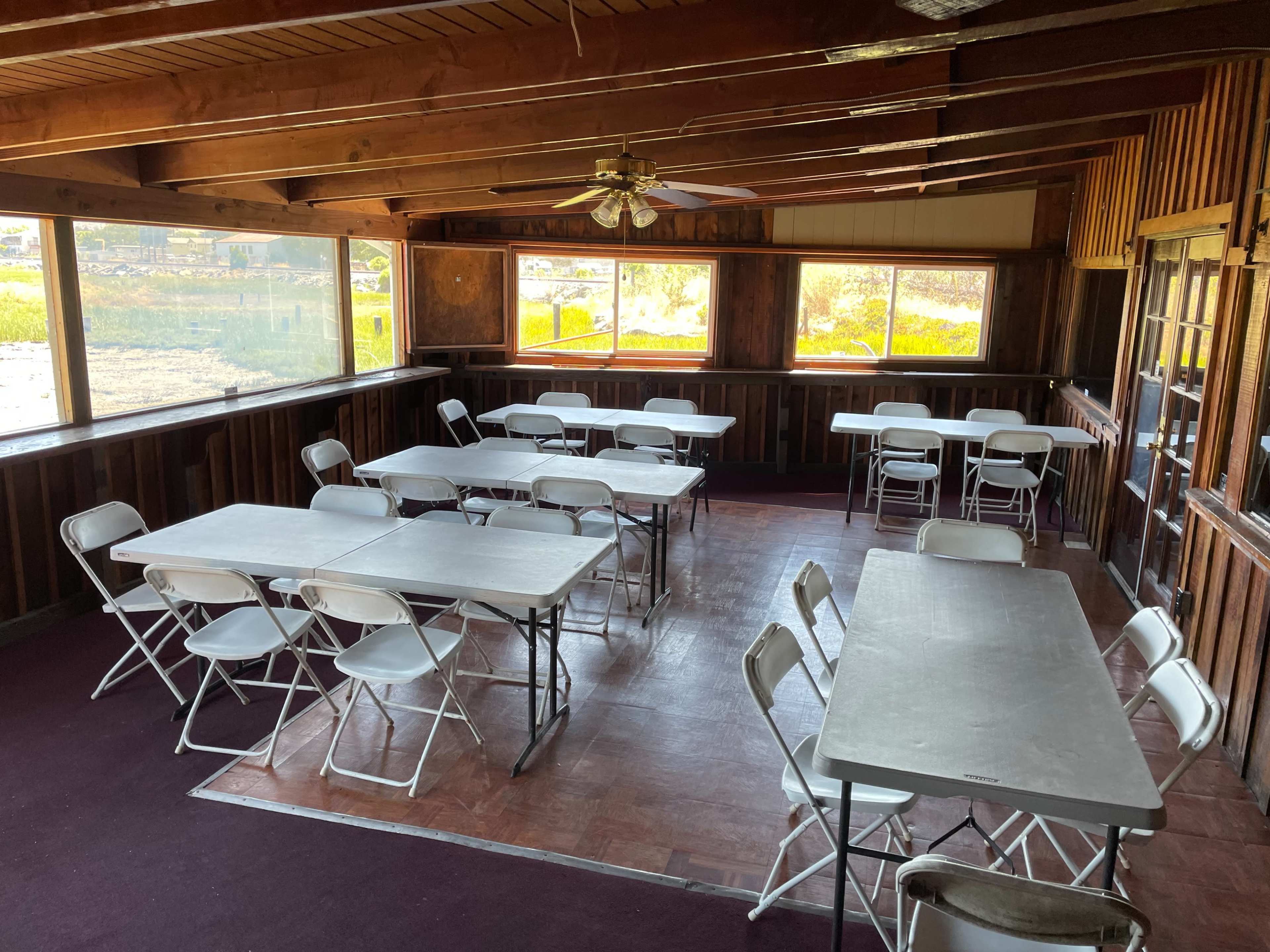 An empty room with wooden walls and a ceiling fan, furnished with several folding tables and chairs arranged for use.