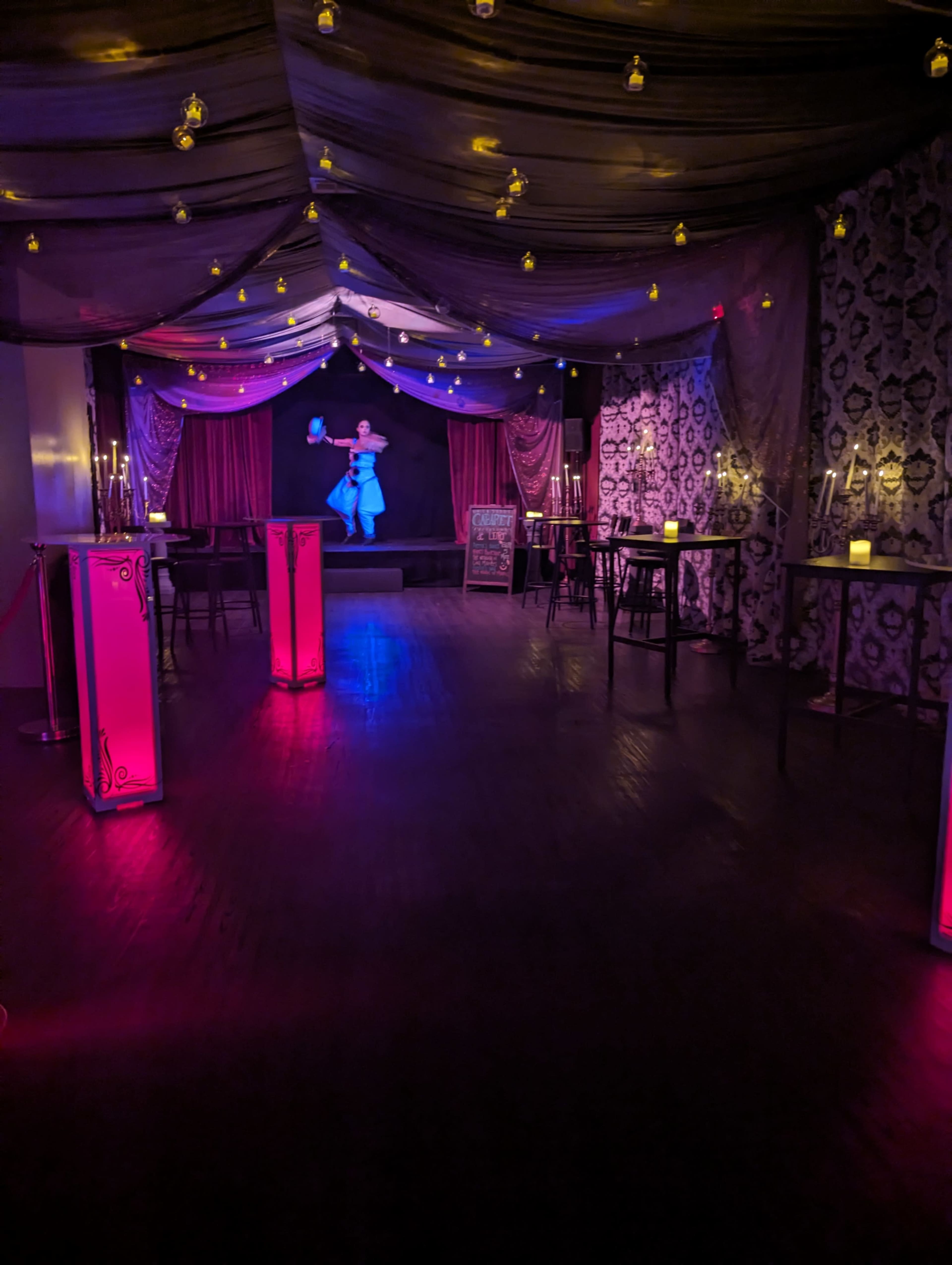 The image shows a dimly lit event space with draped fabric overhead, glowing candles on tables, and a performer in a blue outfit on stage.