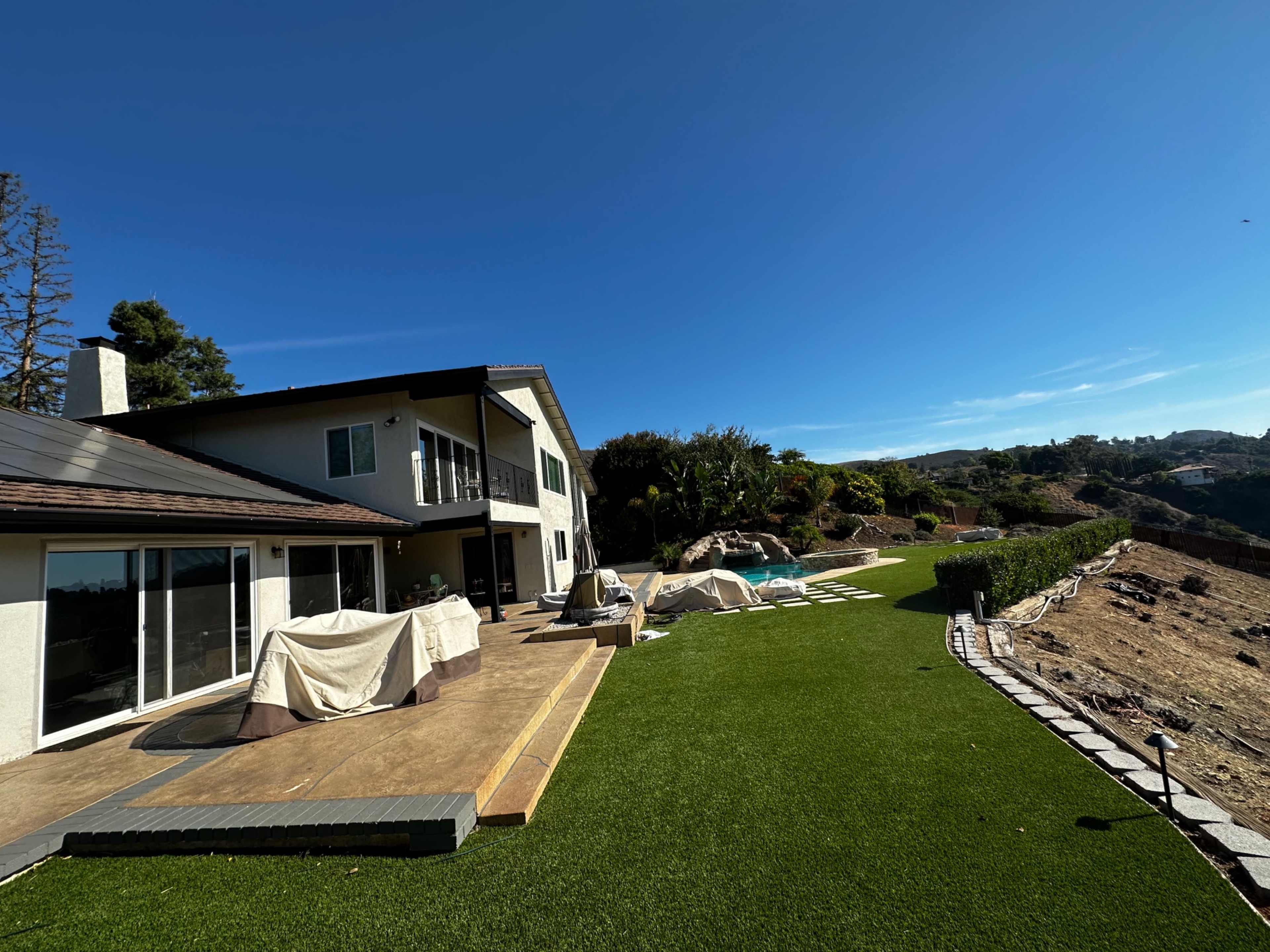 A modern house with a large patio and pool is situated on a hillside surrounded by landscaped lawns and vegetation.