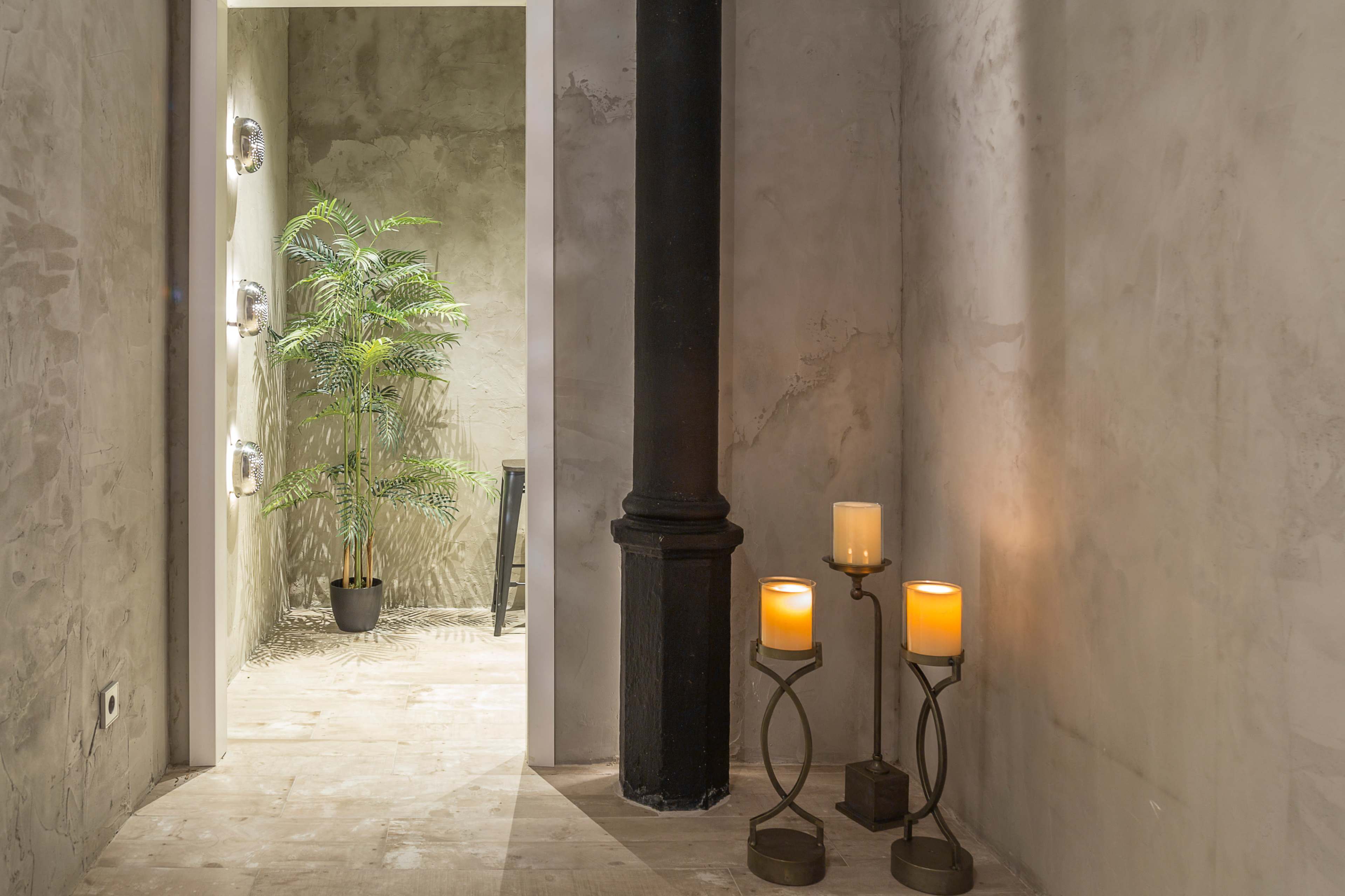 A narrow hallway features a plant in a pot at one end and three candles on a stand near a black column.