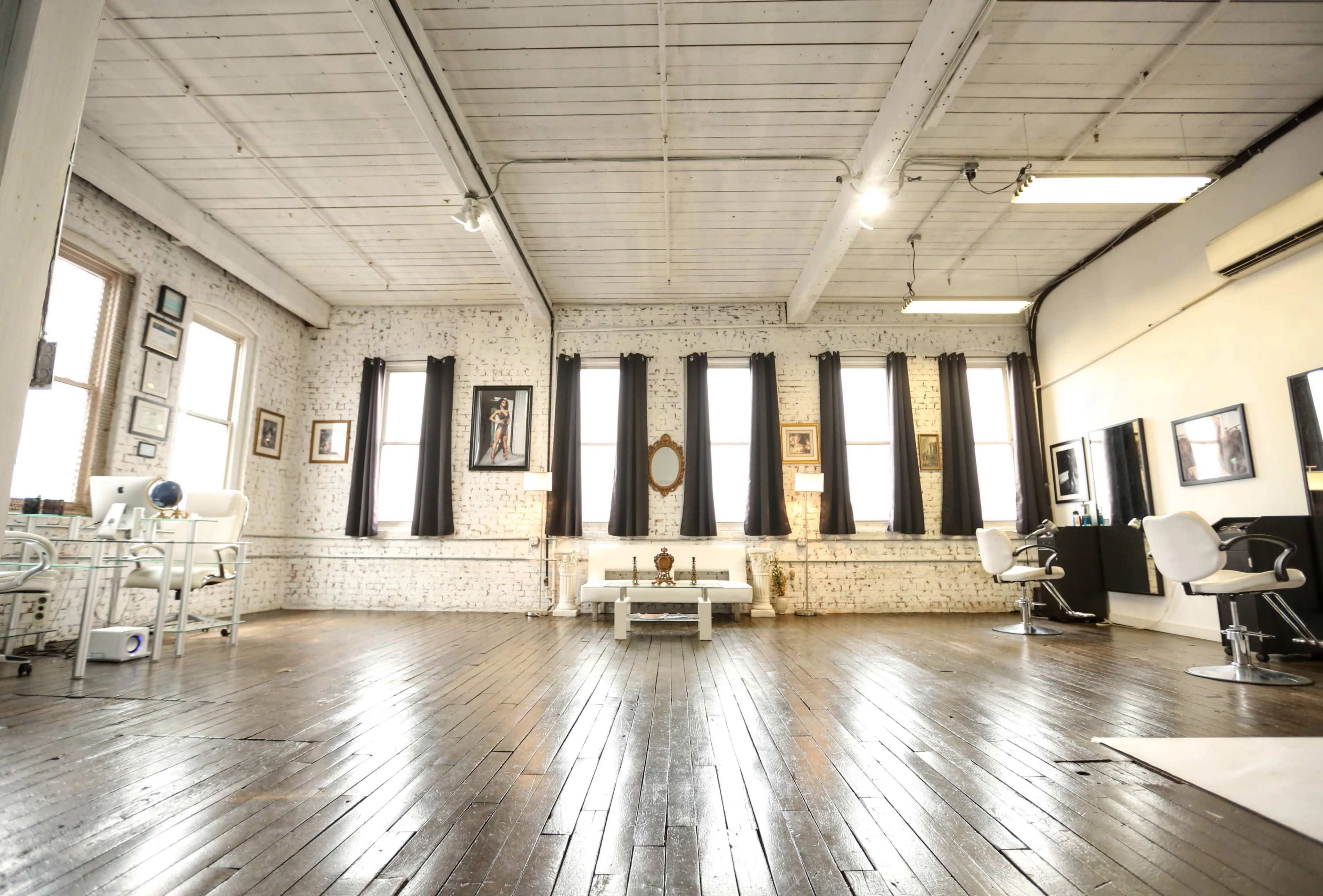 A spacious room with wooden floors, tall windows covered by black curtains, and minimalist furnishings, including two salon chairs and a small coffee table.