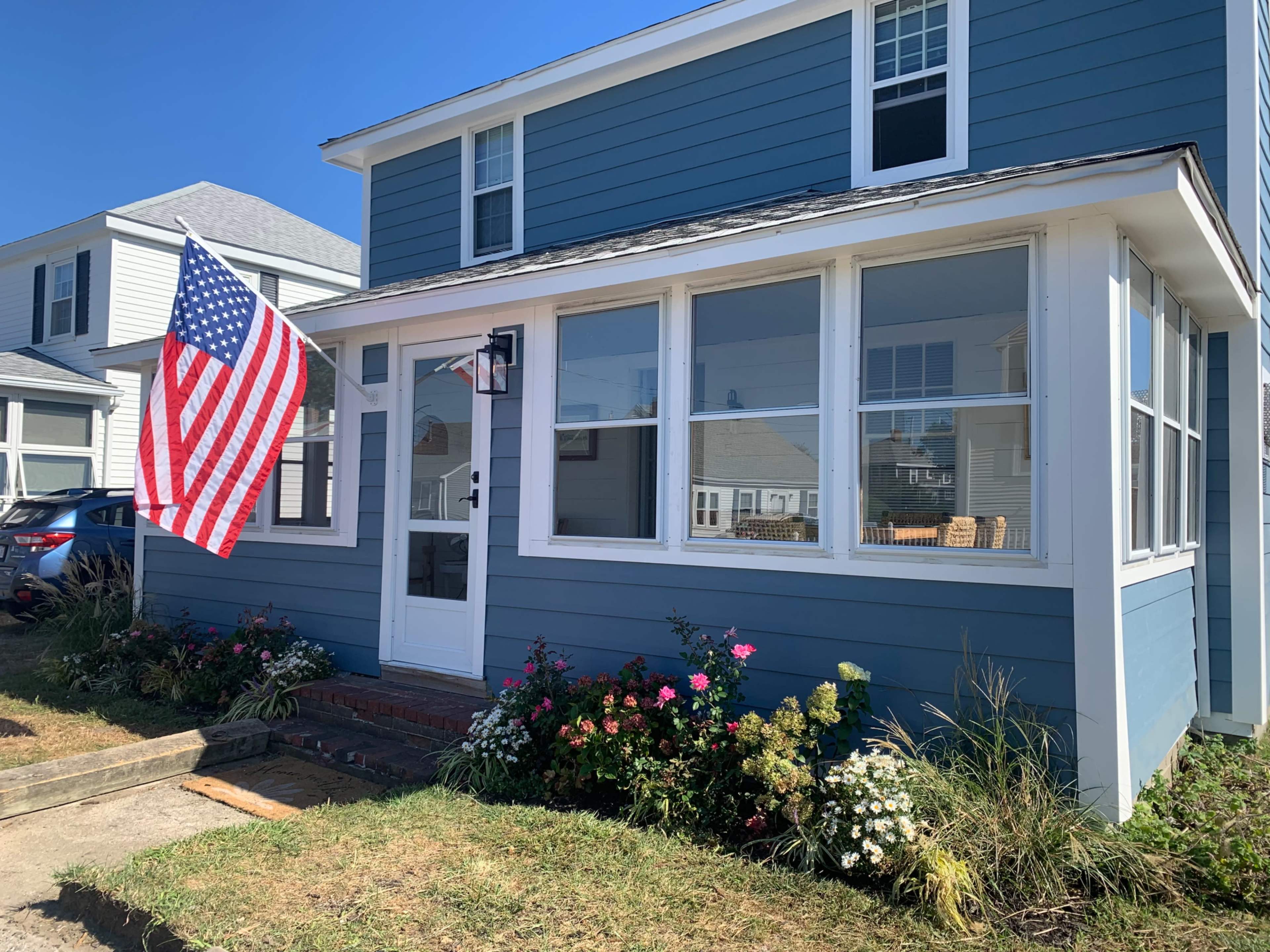 A blue house with white trim features a large American flag hanging from the front porch, surrounded by colorful flowerbeds.