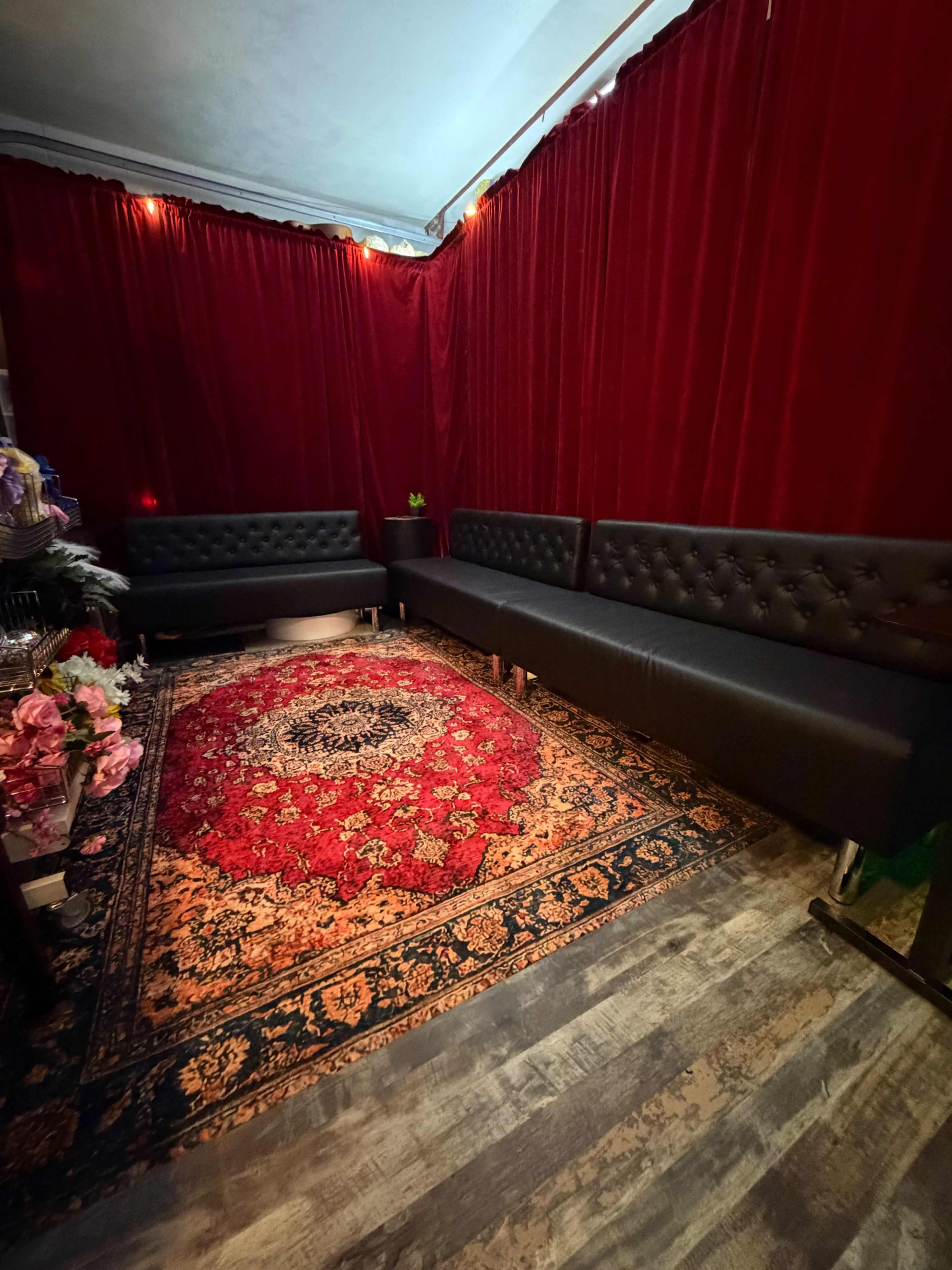 The image shows a cozy seating area with black tufted benches against a backdrop of red curtains and a large, ornate red and black rug on the floor.