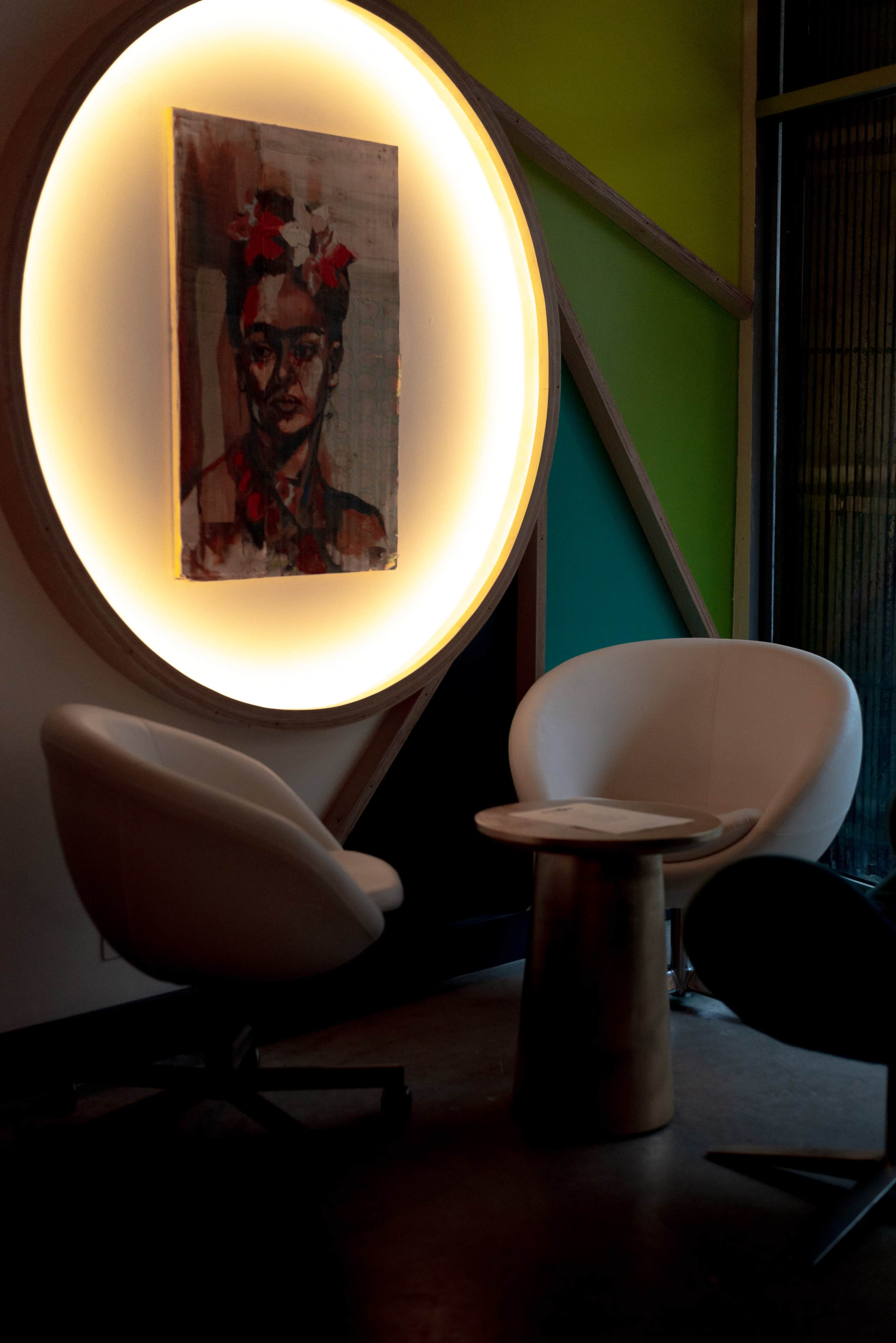 Two white chairs and a round table positioned beneath a lit circular frame displaying a portrait of a woman.
