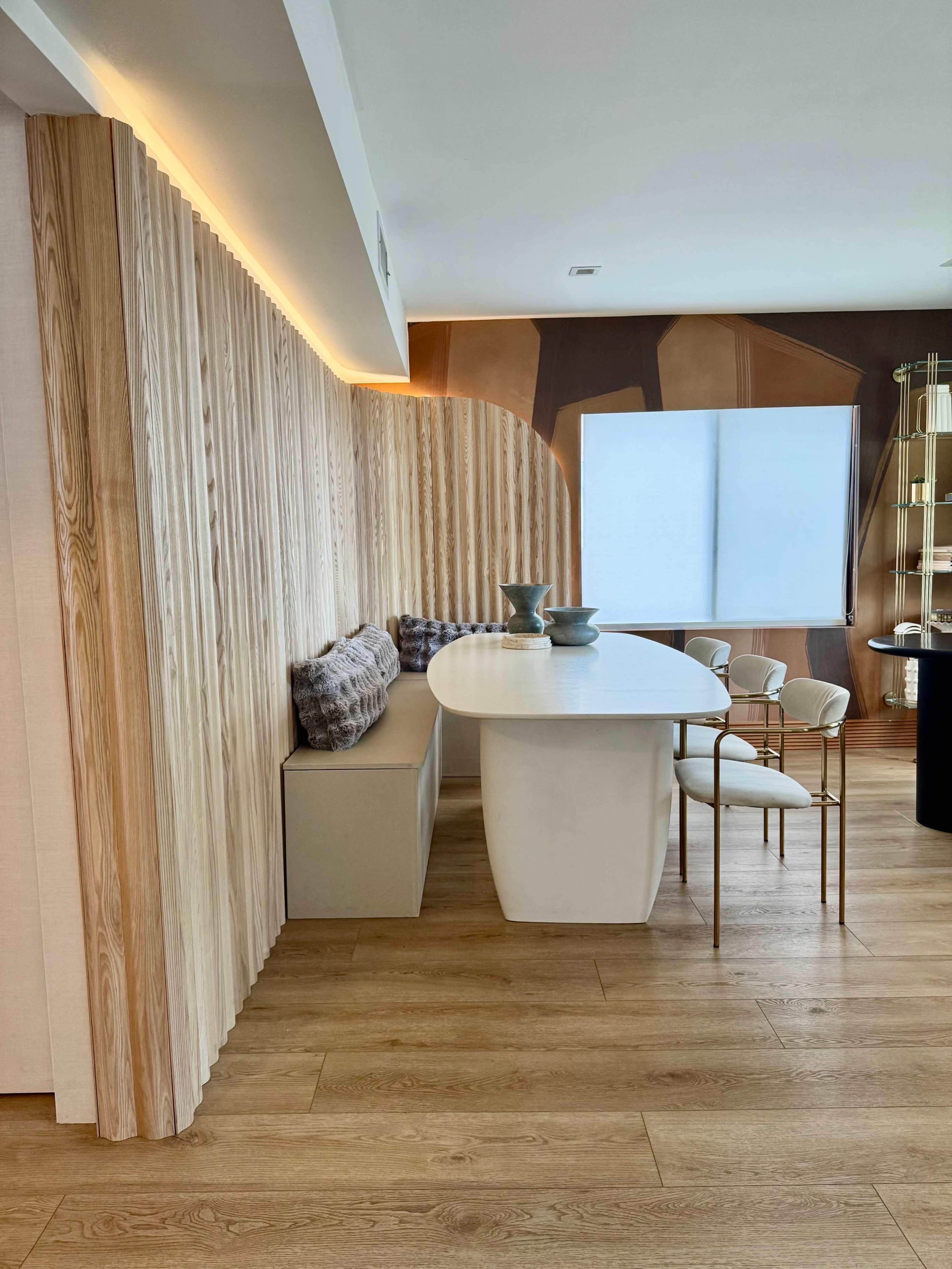 The image shows a modern dining area featuring a round white table, beige seating, and wooden wall paneling, illuminated by soft lighting.
