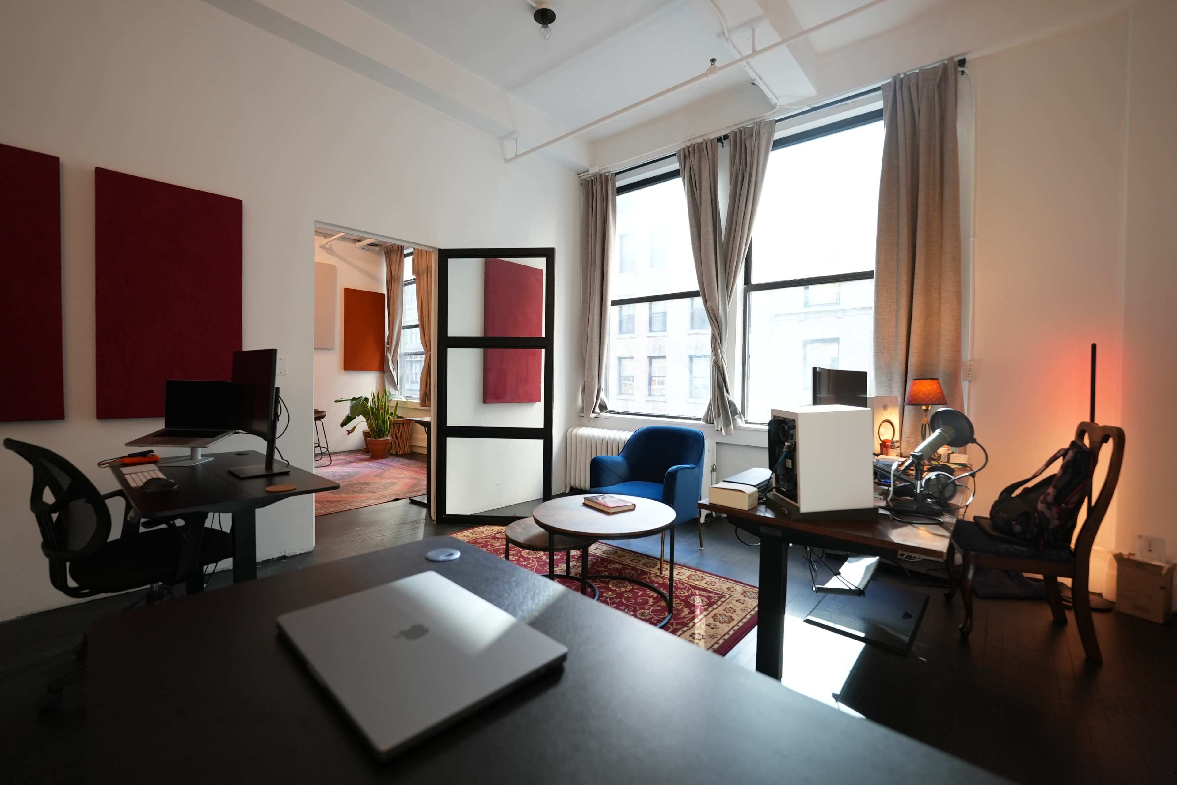 A bright office space featuring two workstations, a blue chair, a coffee table, and large windows with curtains, along with a doorway leading to another room.