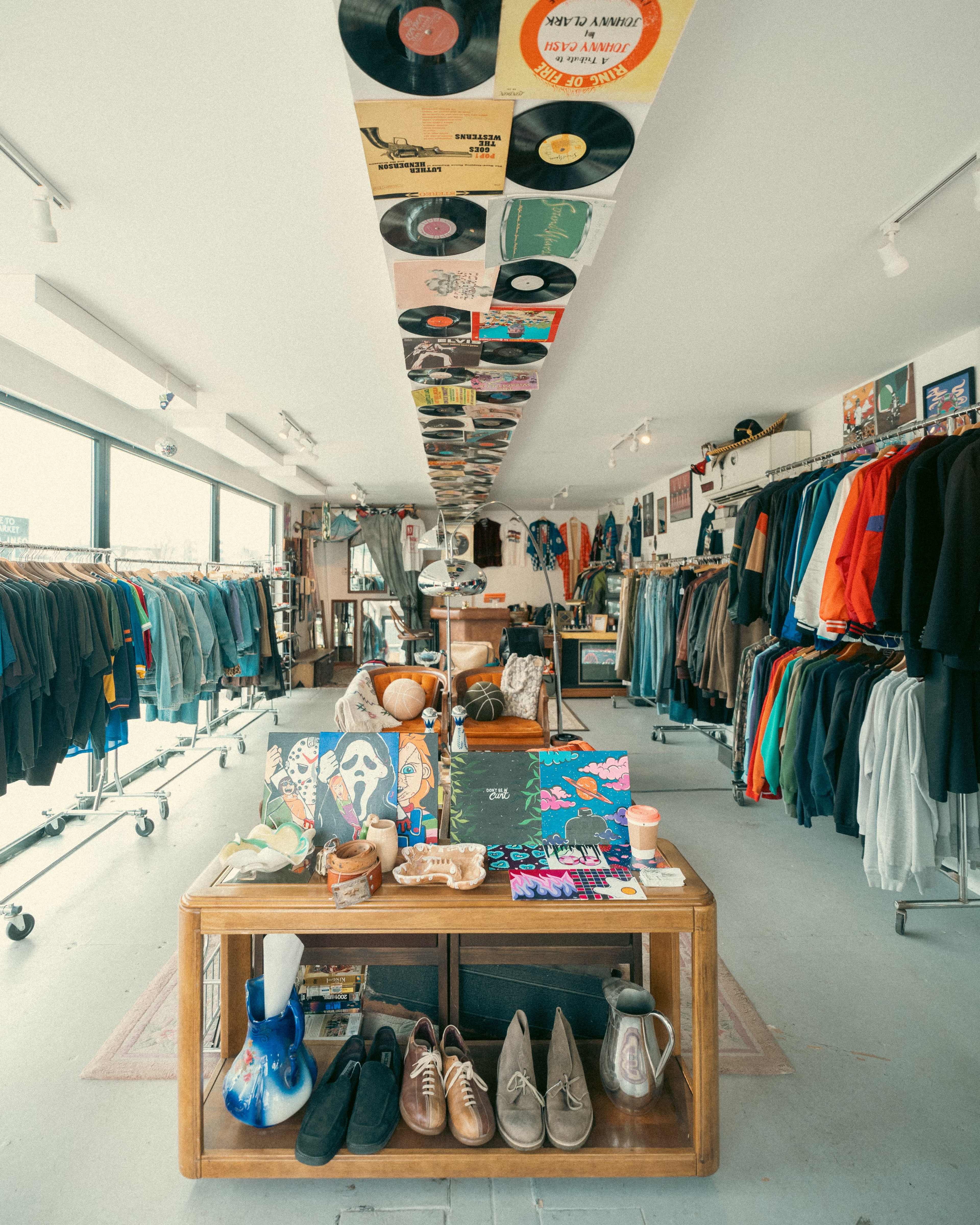 The image shows a vintage clothing store featuring a long display table with various items, surrounded by clothing racks and a ceiling adorned with vinyl records.
