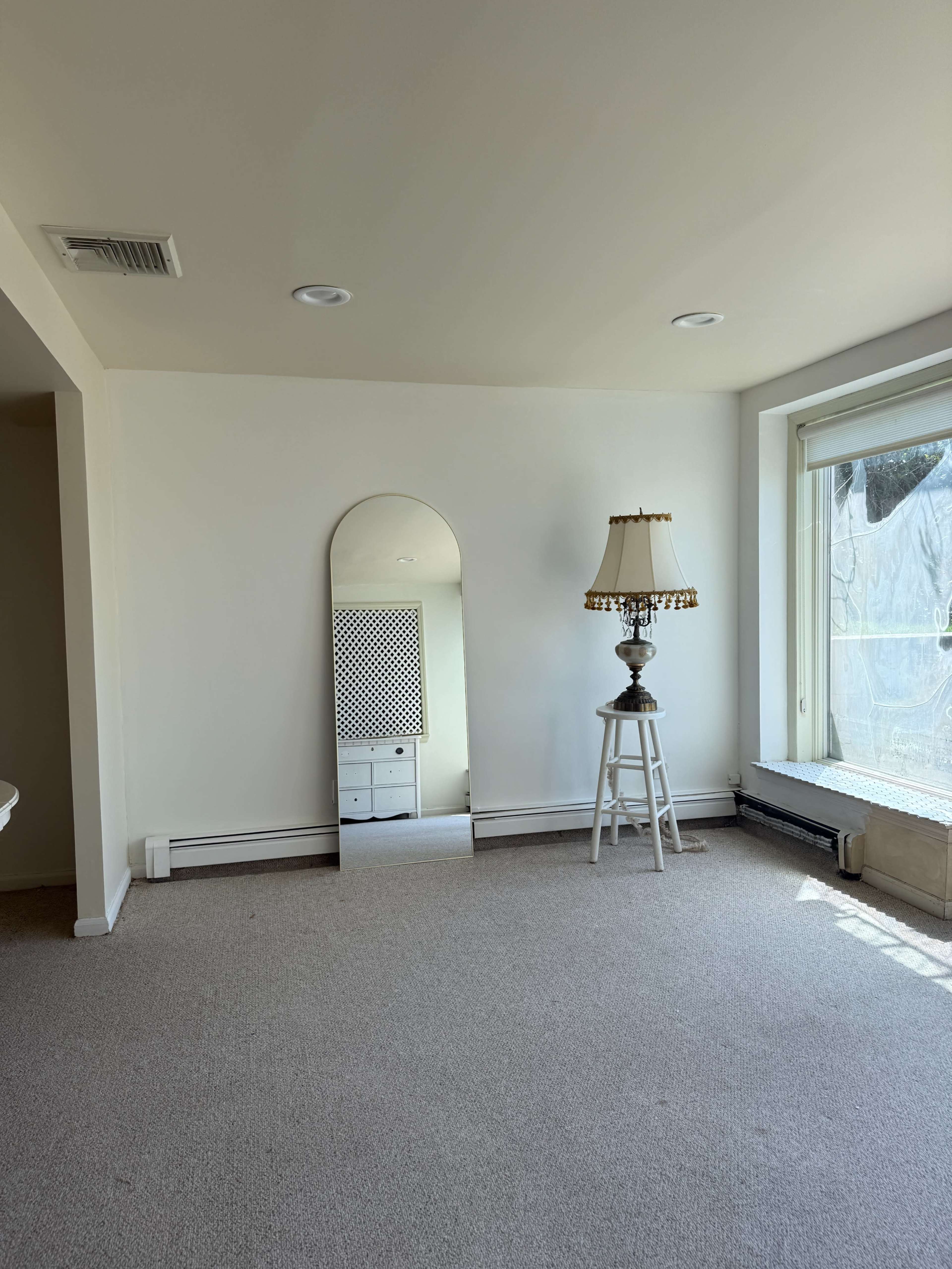 The image shows a sparsely furnished room with beige carpet, a large mirror leaning against a wall, a lamp on a small table, and a window with partially drawn curtains.