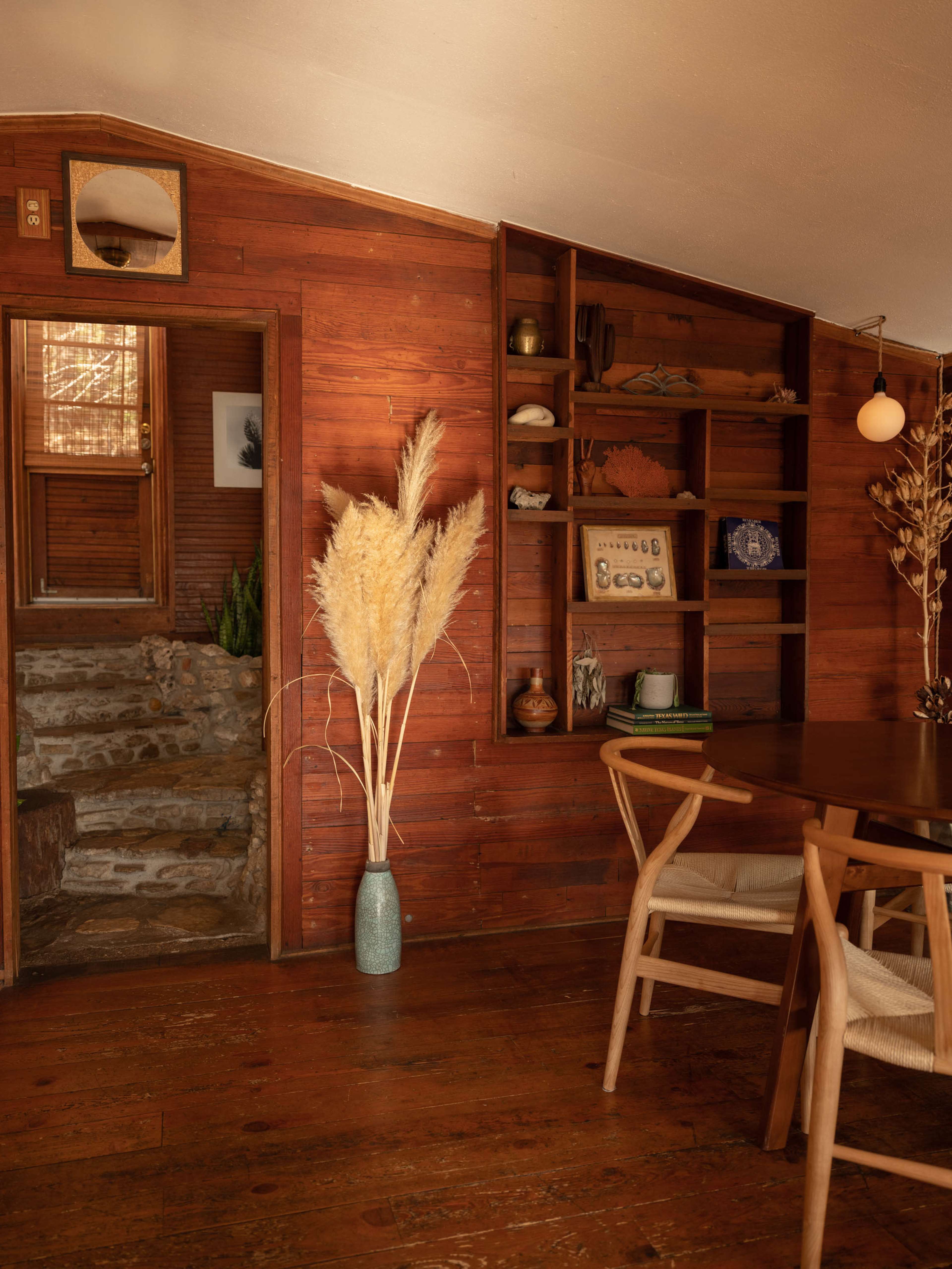 A cozy interior features wooden walls, a stone staircase, a vase of pampas grass, and a dining table with wooden chairs.