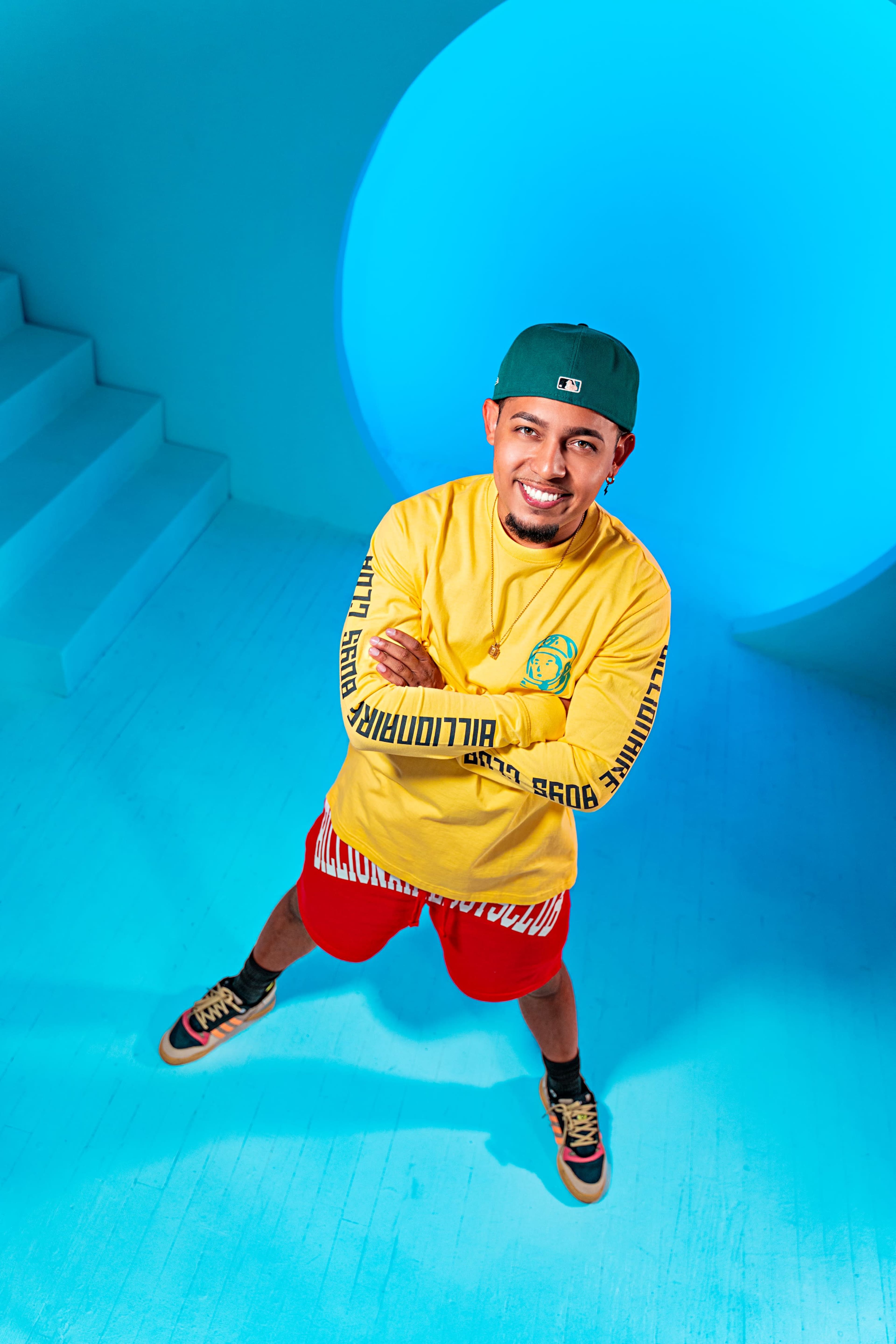 A young man in a yellow long-sleeve shirt and red shorts stands confidently on a light blue floor, with a circular blue backdrop and steps behind him.
