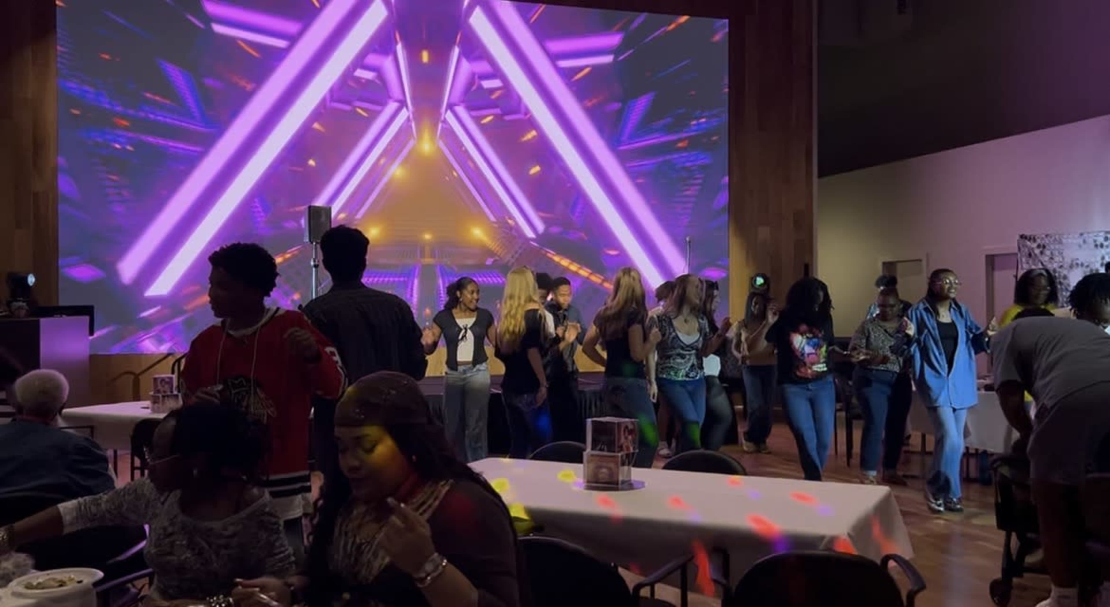 A group of people dances in front of a vibrant, illuminated backdrop with colorful geometric patterns at an event venue.