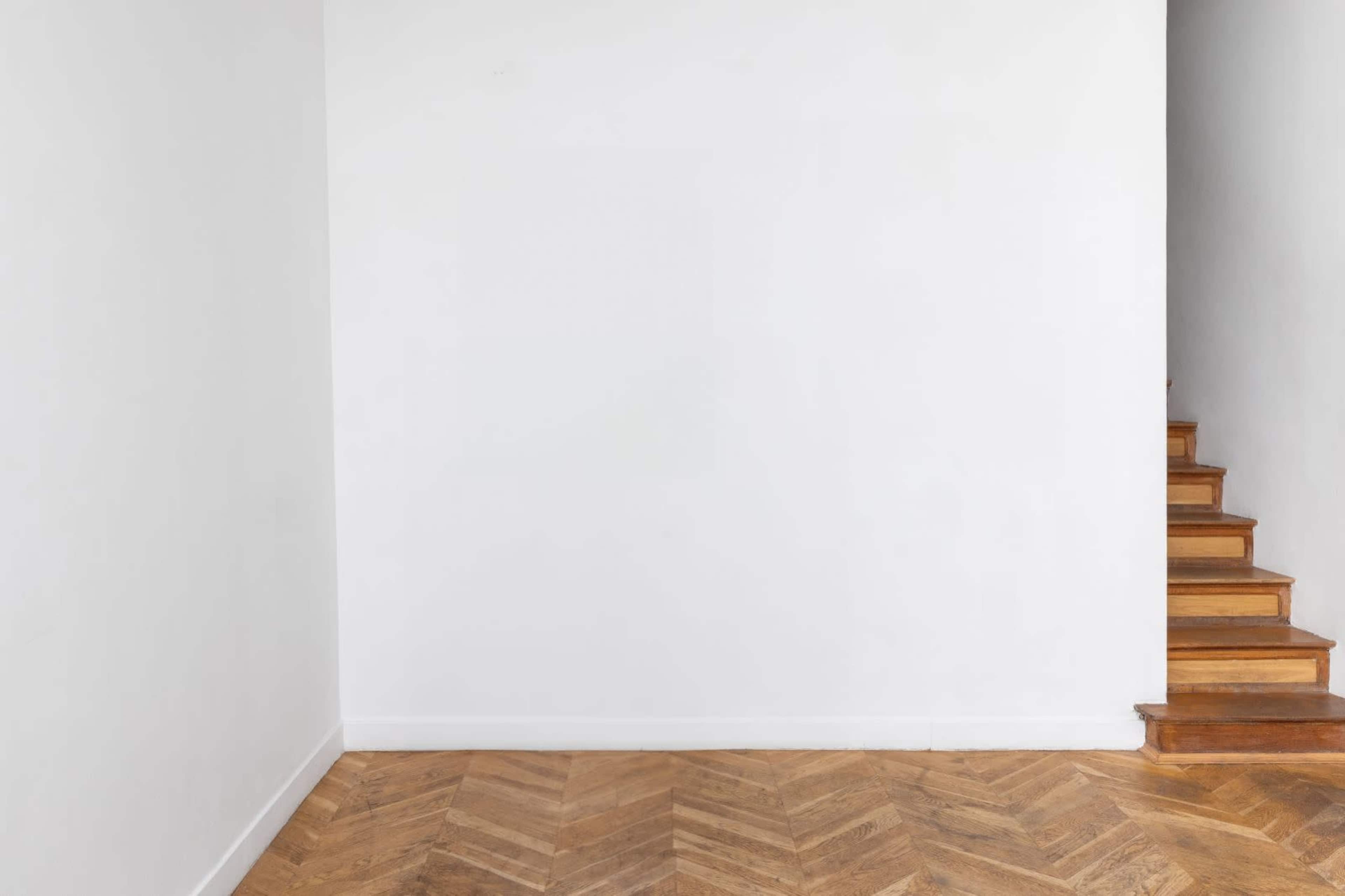 The image shows a minimalistic interior with a white wall, wooden herringbone flooring, and a staircase in the background.