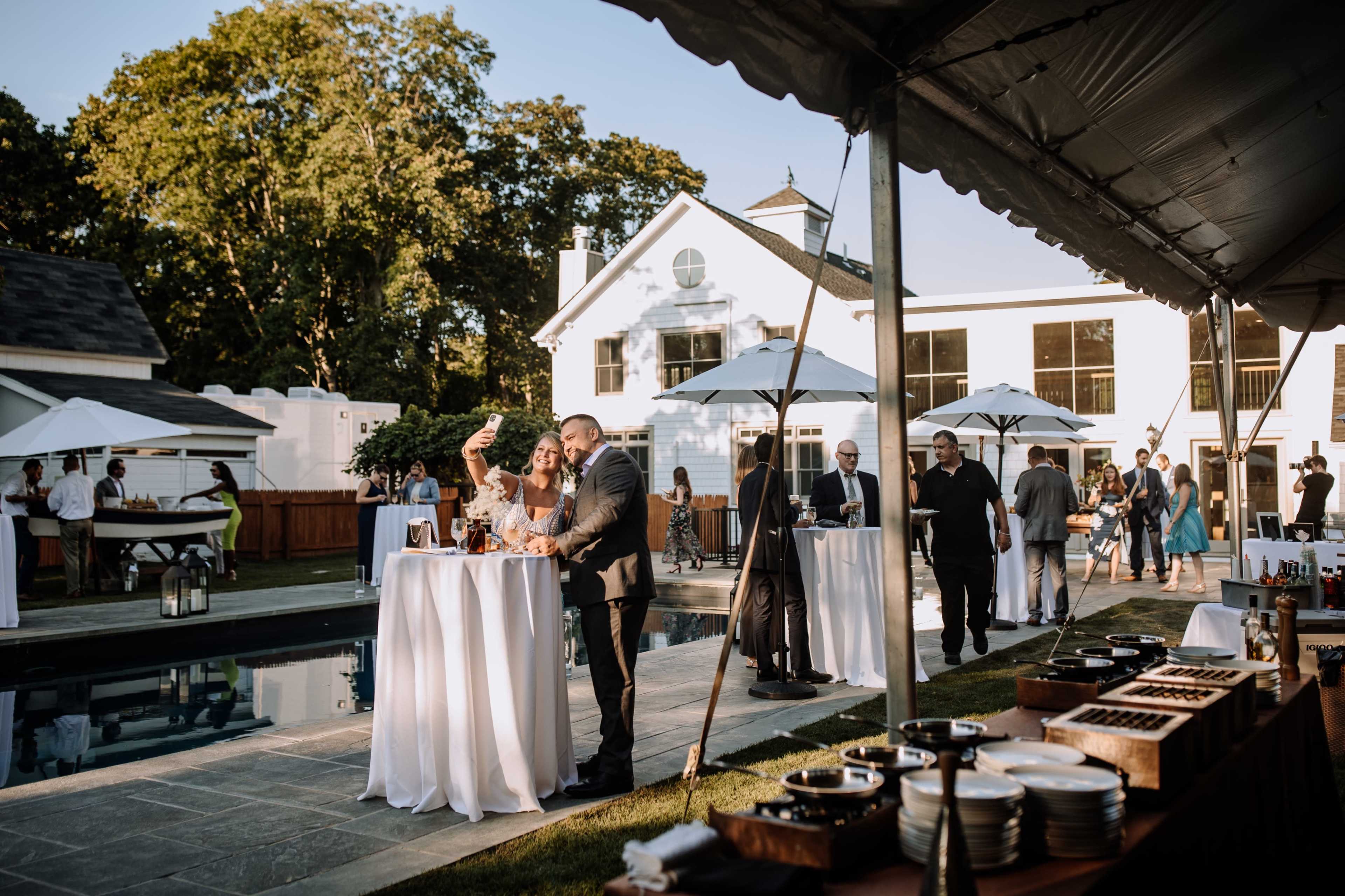 A couple takes a selfie near a pool while guests enjoy a reception under white tents in a landscaped outdoor setting.