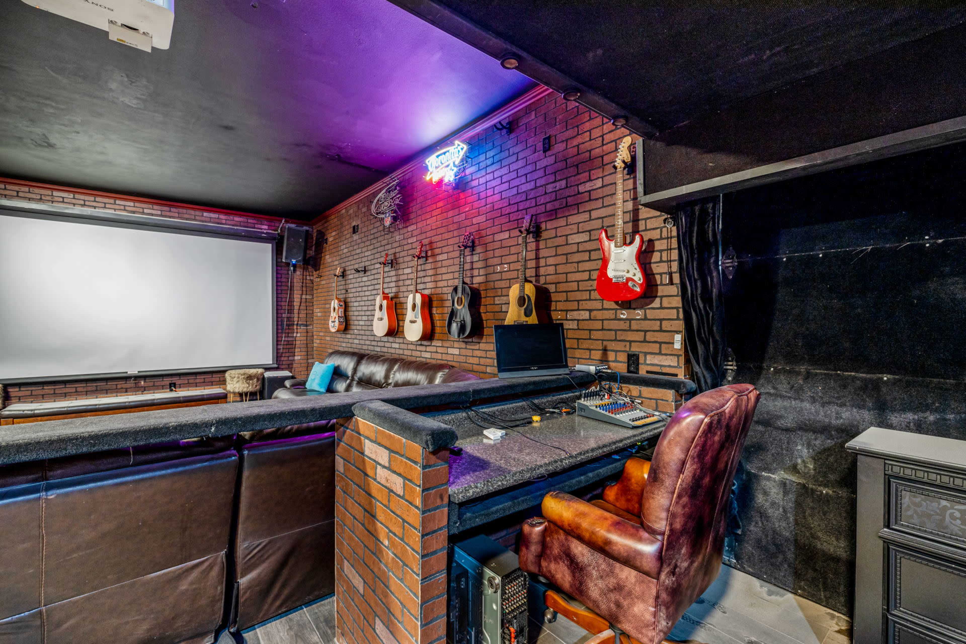 A dimly lit room features a soundboard, leather seating, and a wall adorned with various guitars against a brick backdrop.