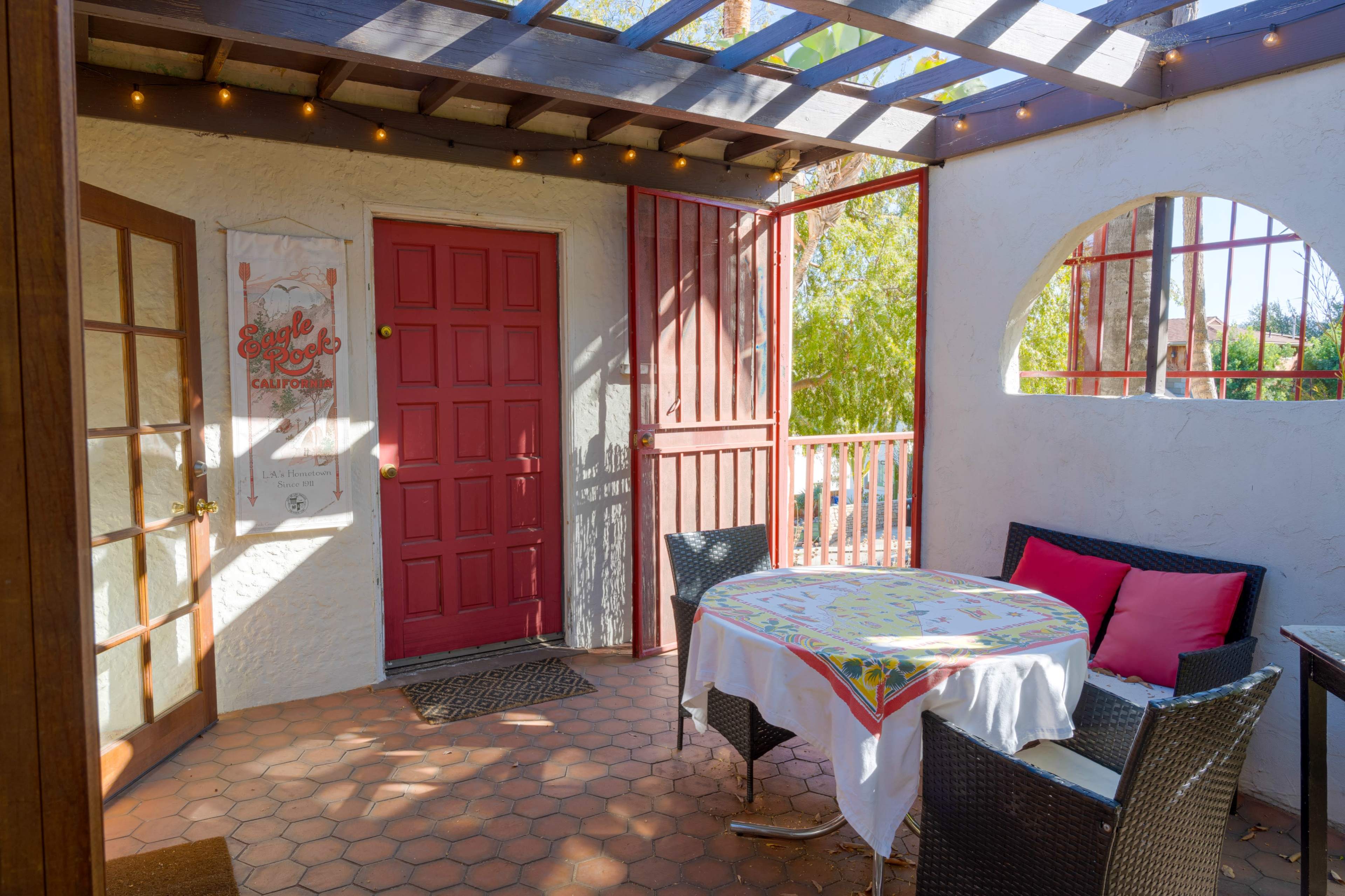 A patio area features a round table with a tablecloth and several chairs, surrounded by a red door and large windows allowing natural light to enter.