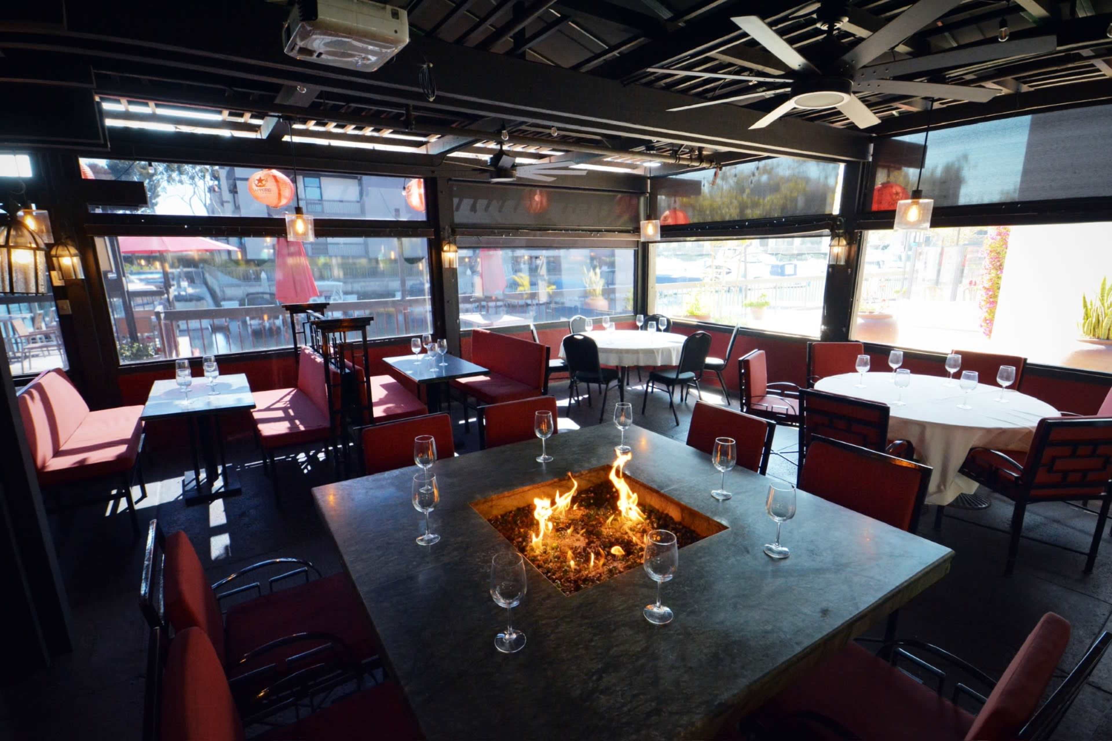 A modern restaurant interior featuring round and rectangular tables with red seating, a central fire pit, and large windows allowing natural light.