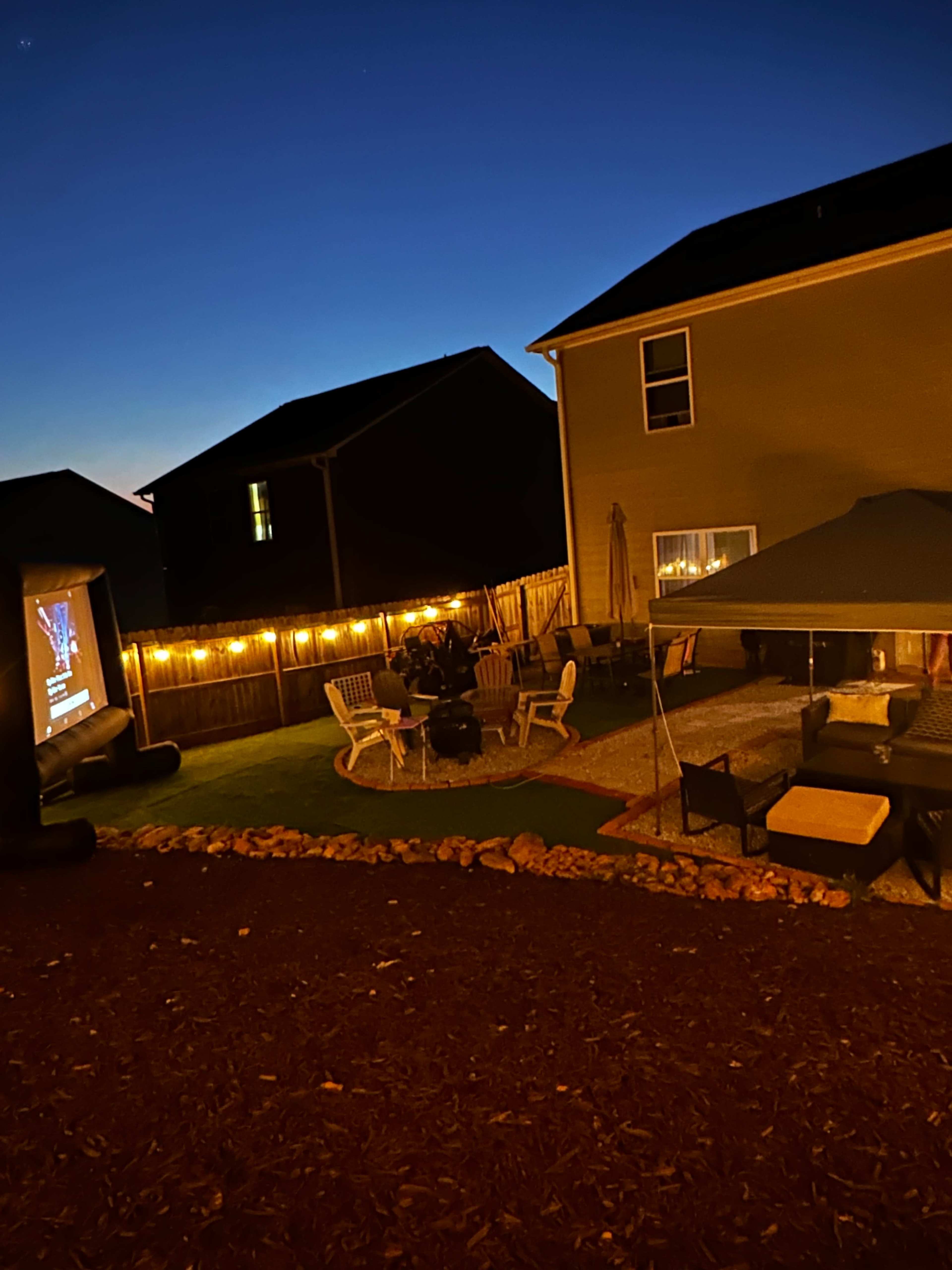 A backyard is illuminated by string lights, featuring seating areas and a movie screen set against a twilight sky.