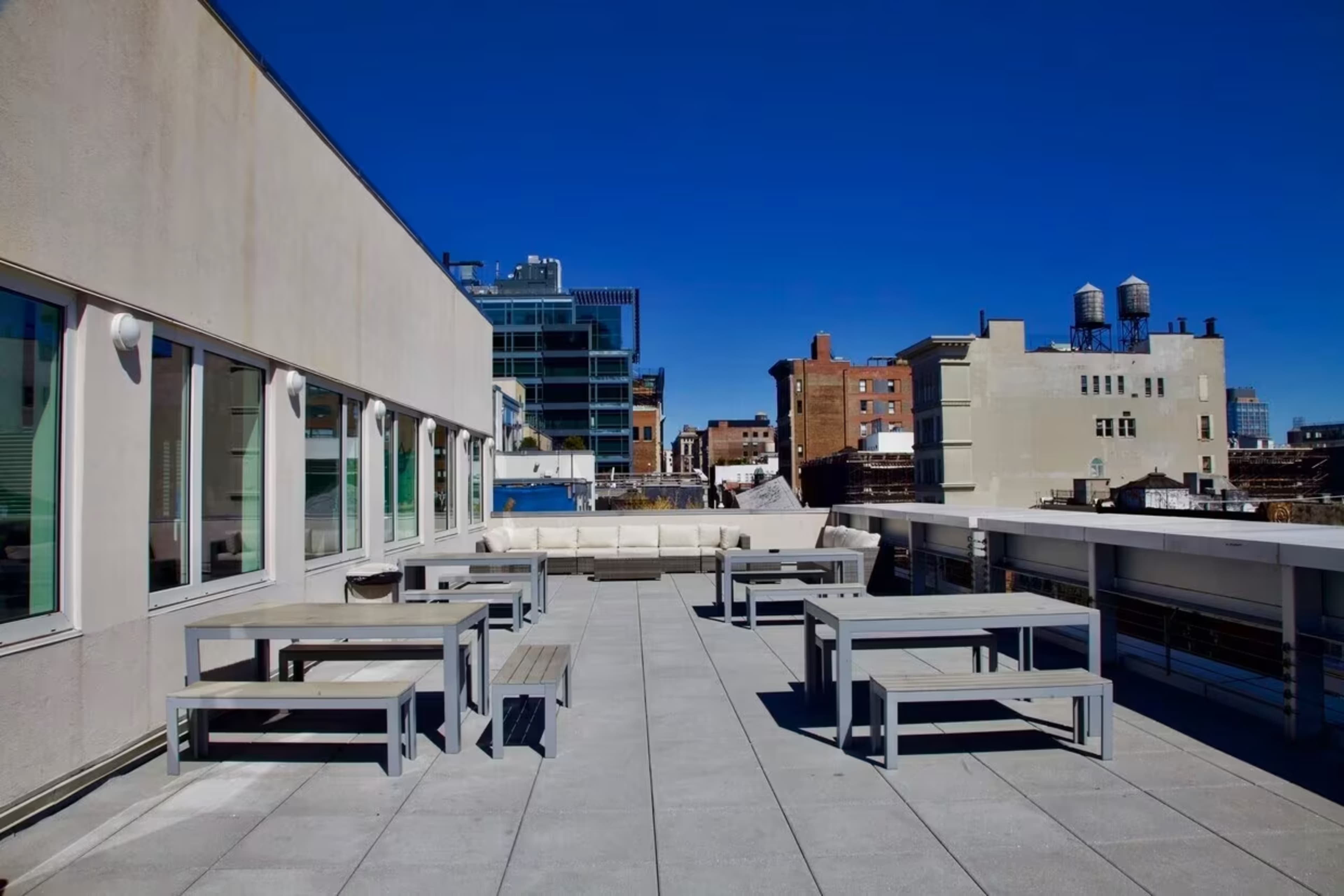 A rooftop terrace featuring several gray tables and benches under a clear blue sky, with buildings partially visible in the background.