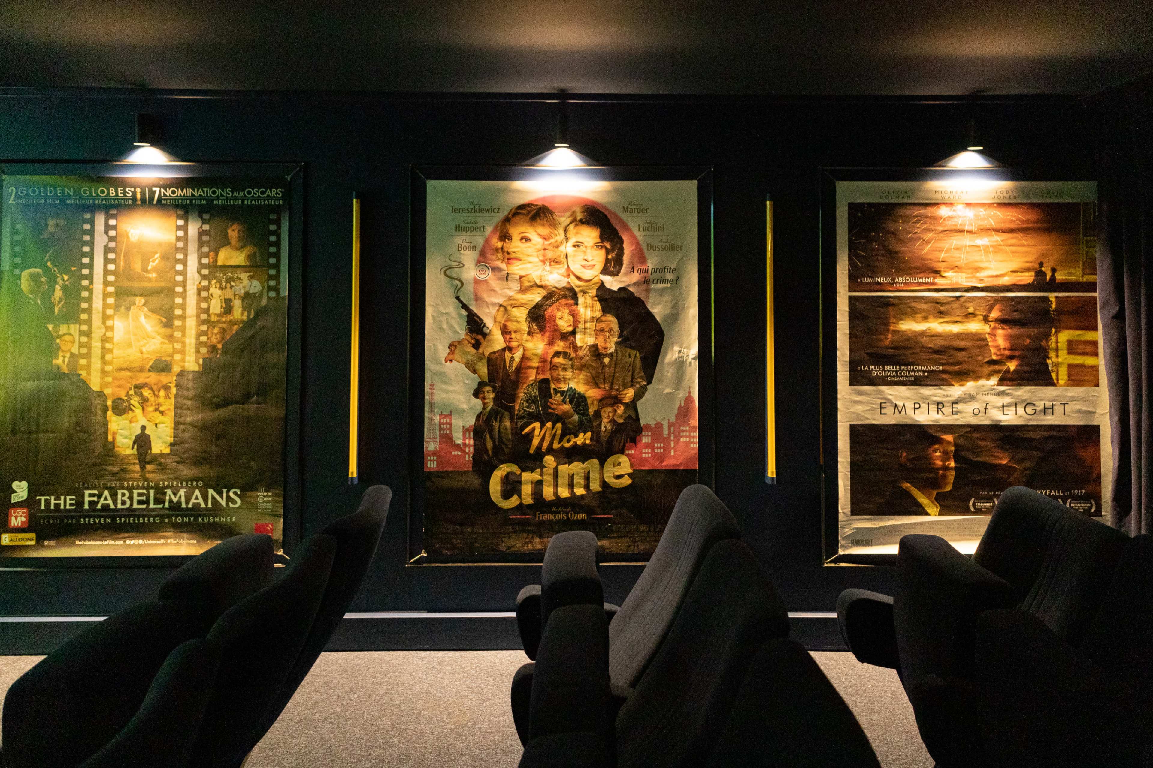 A dimly lit movie theater with three framed film posters displayed on the wall, alongside several empty, dark-colored chairs in front of them.