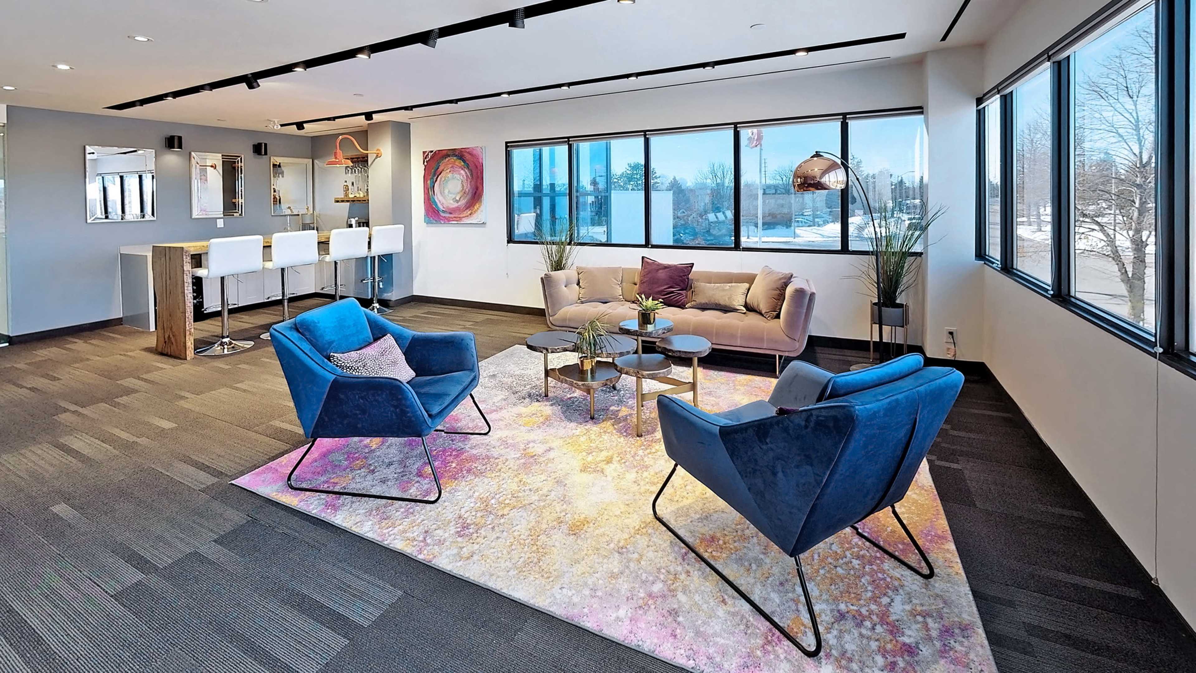The image shows a modern office lounge area with two blue chairs, a gray sofa, and a coffee table on a colorful rug, set against large windows letting in natural light.