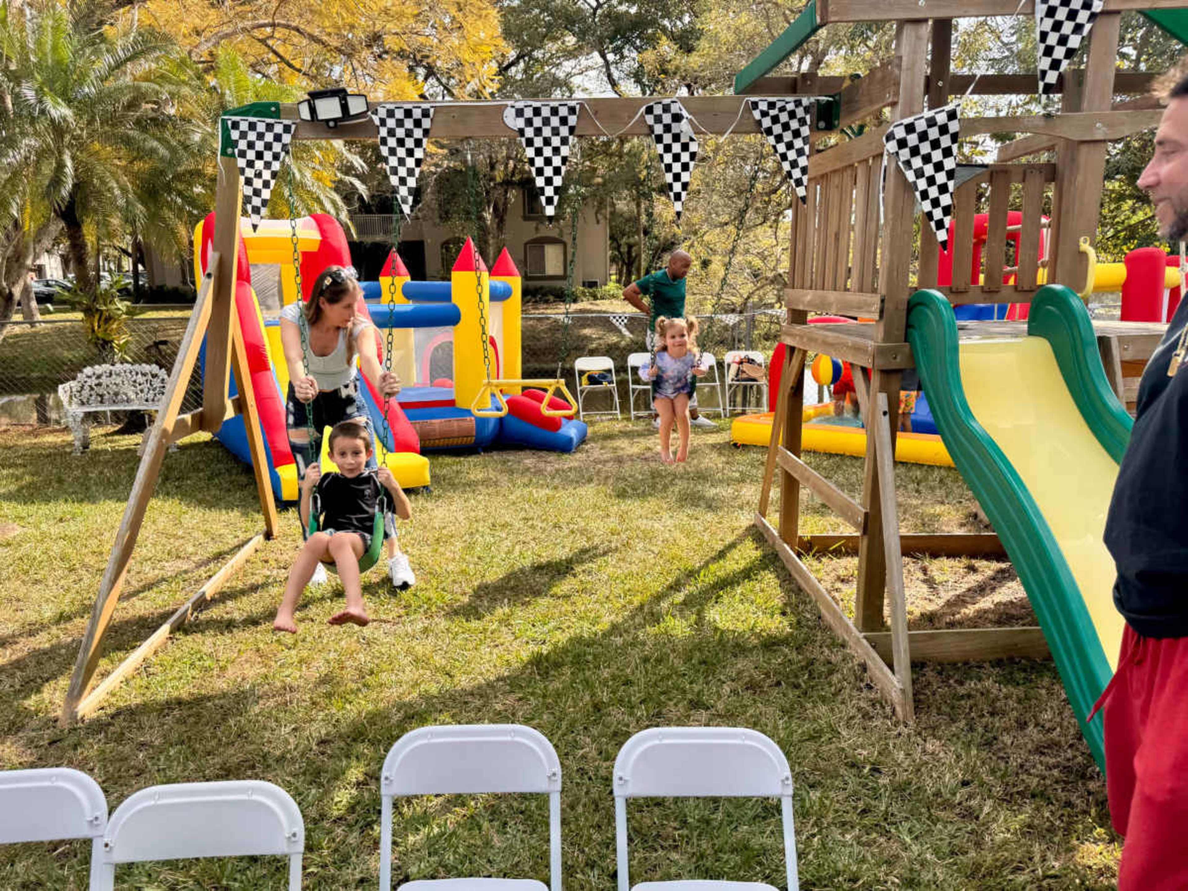 The image shows a colorful outdoor playground area with children playing on swings, slides, and various inflatable structures, surrounded by chairs arranged in a row.