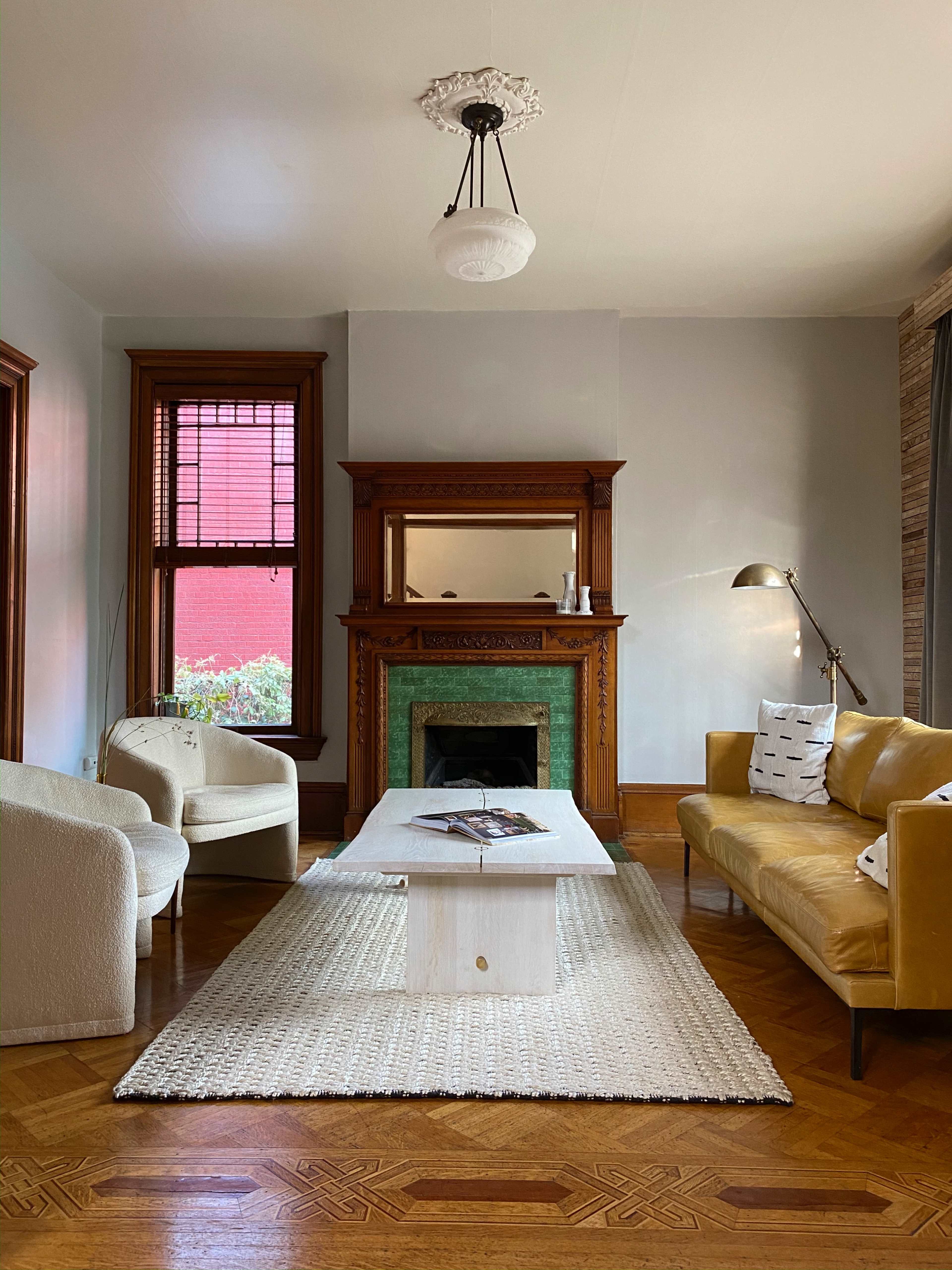 A living room with a yellow sofa, white armchairs, a coffee table on a woven rug, and an ornate fireplace with a mirror above it.
