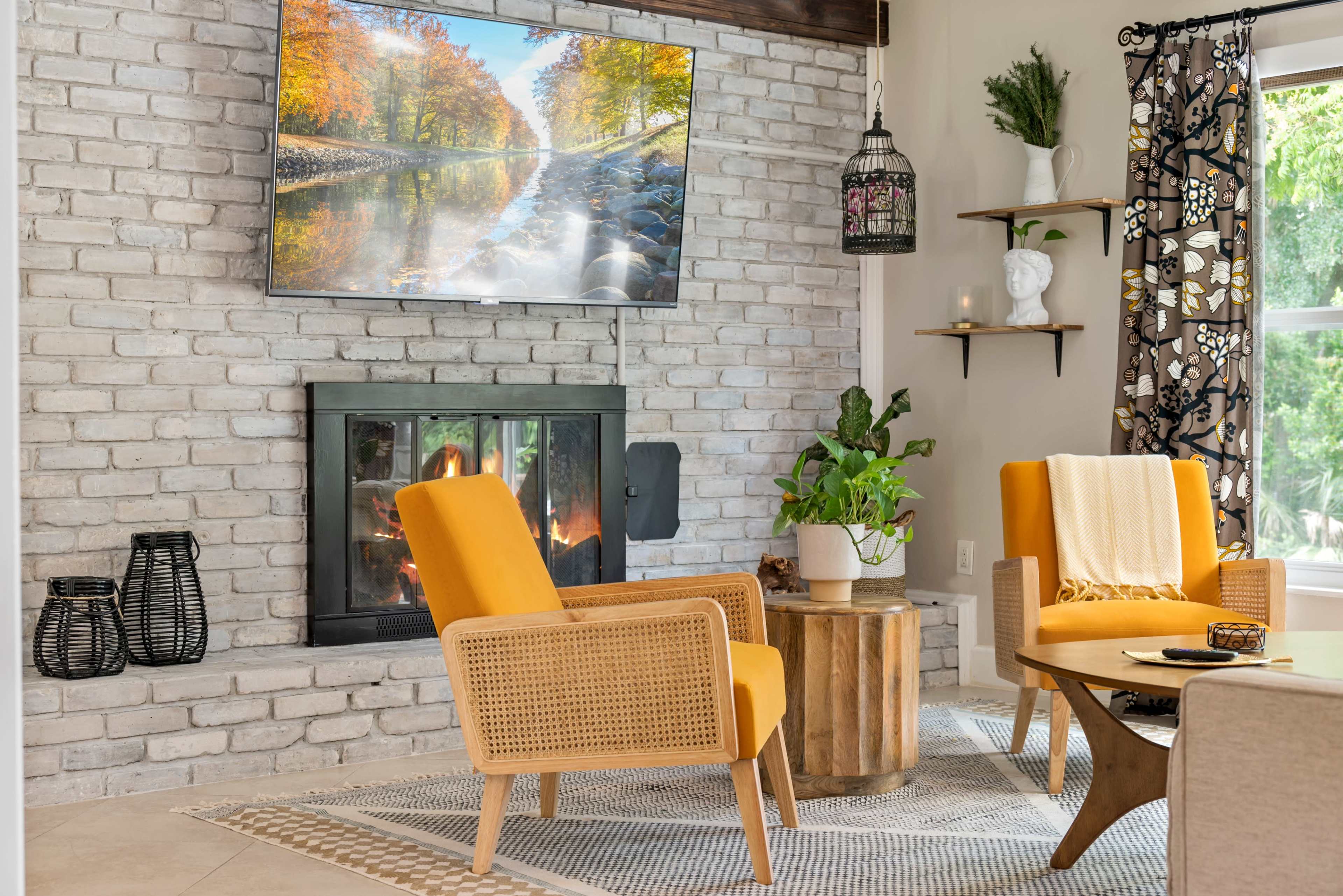 A modern living room features two yellow chairs facing a round wooden table, with a stone fireplace and a television mounted above it.