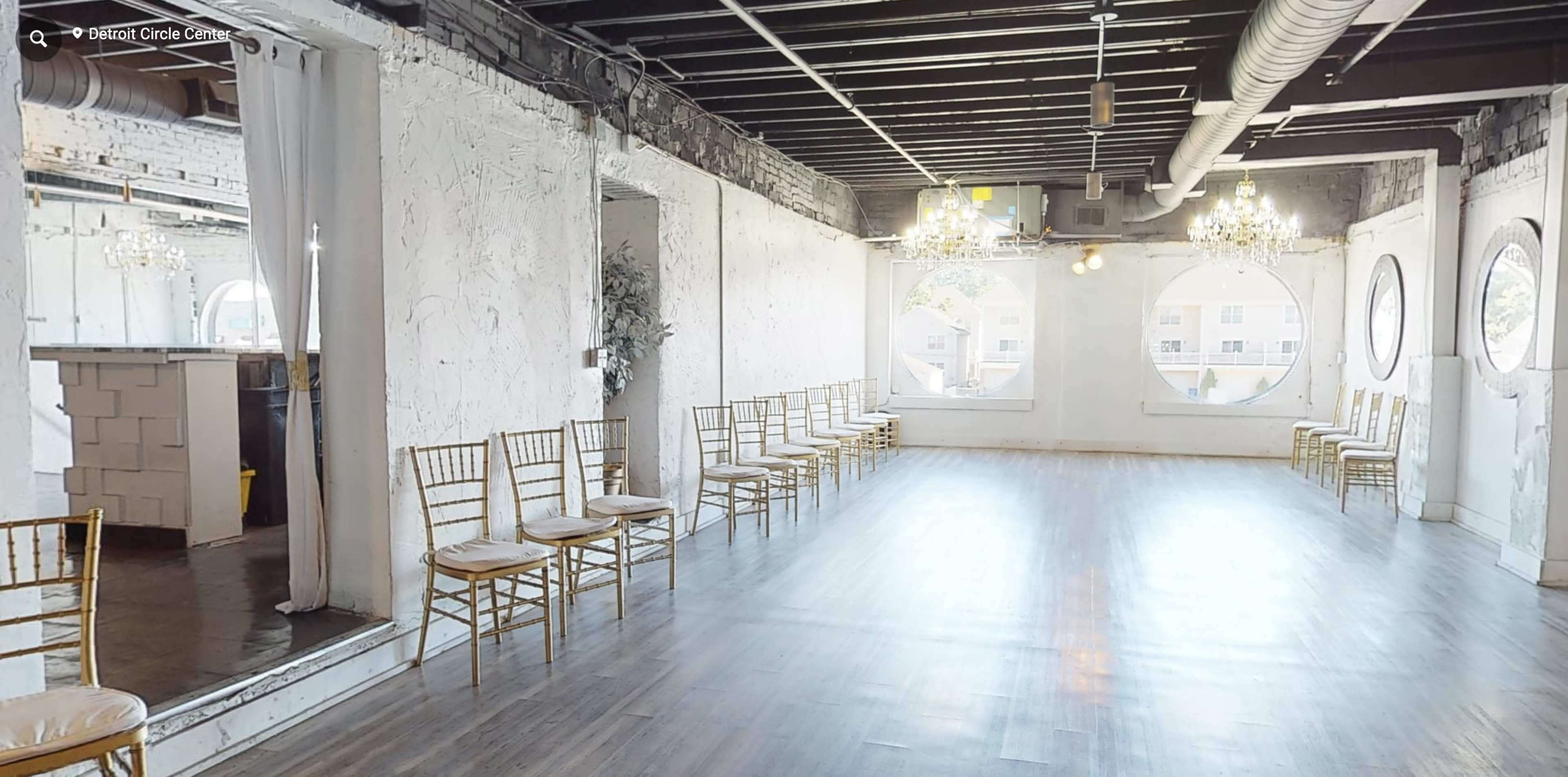 A spacious event room with wooden flooring, round windows, and rows of empty gold chairs along the walls.
