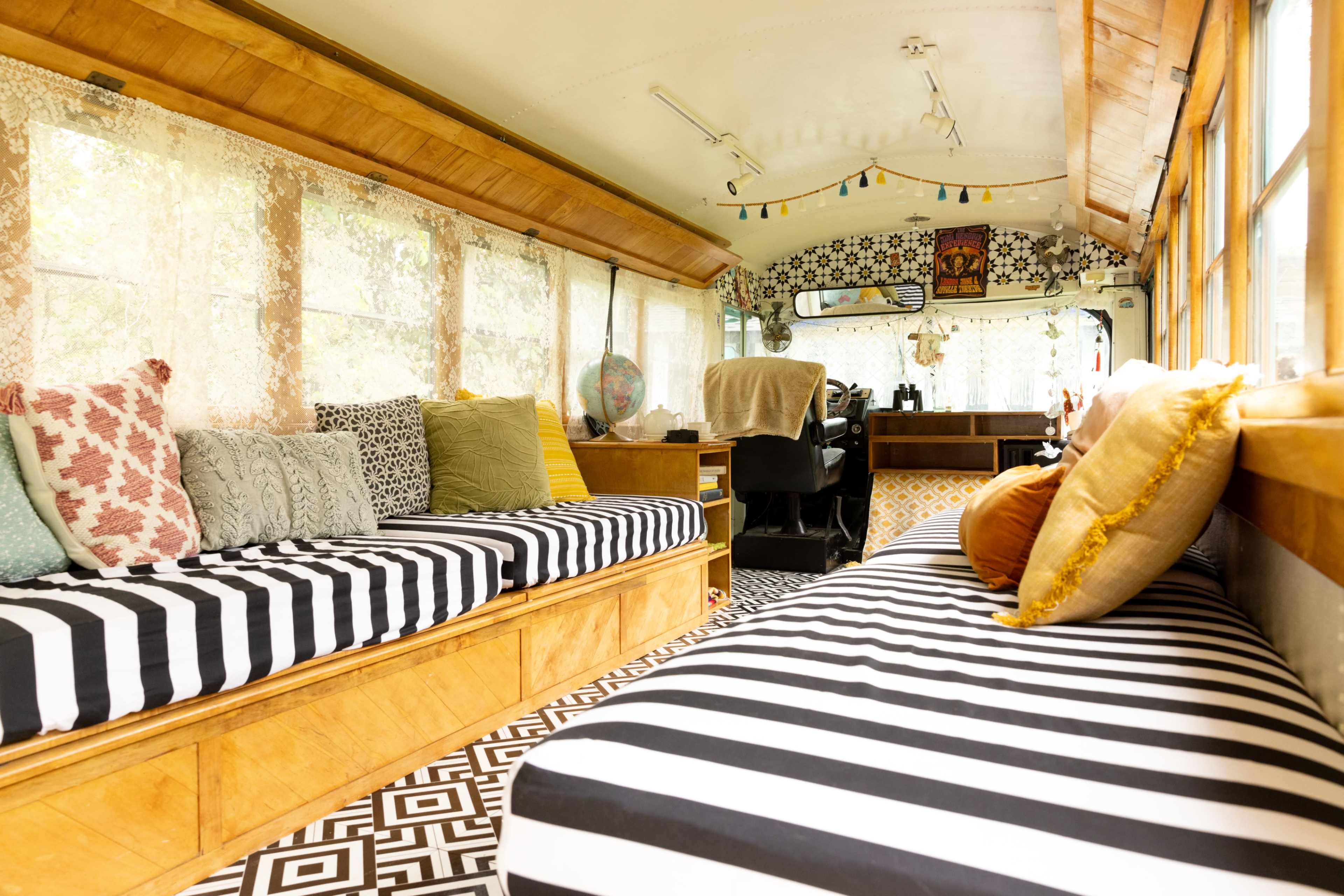 The interior of the converted bus features striped seating, a wooden desk area, and large windows allowing natural light to fill the space.