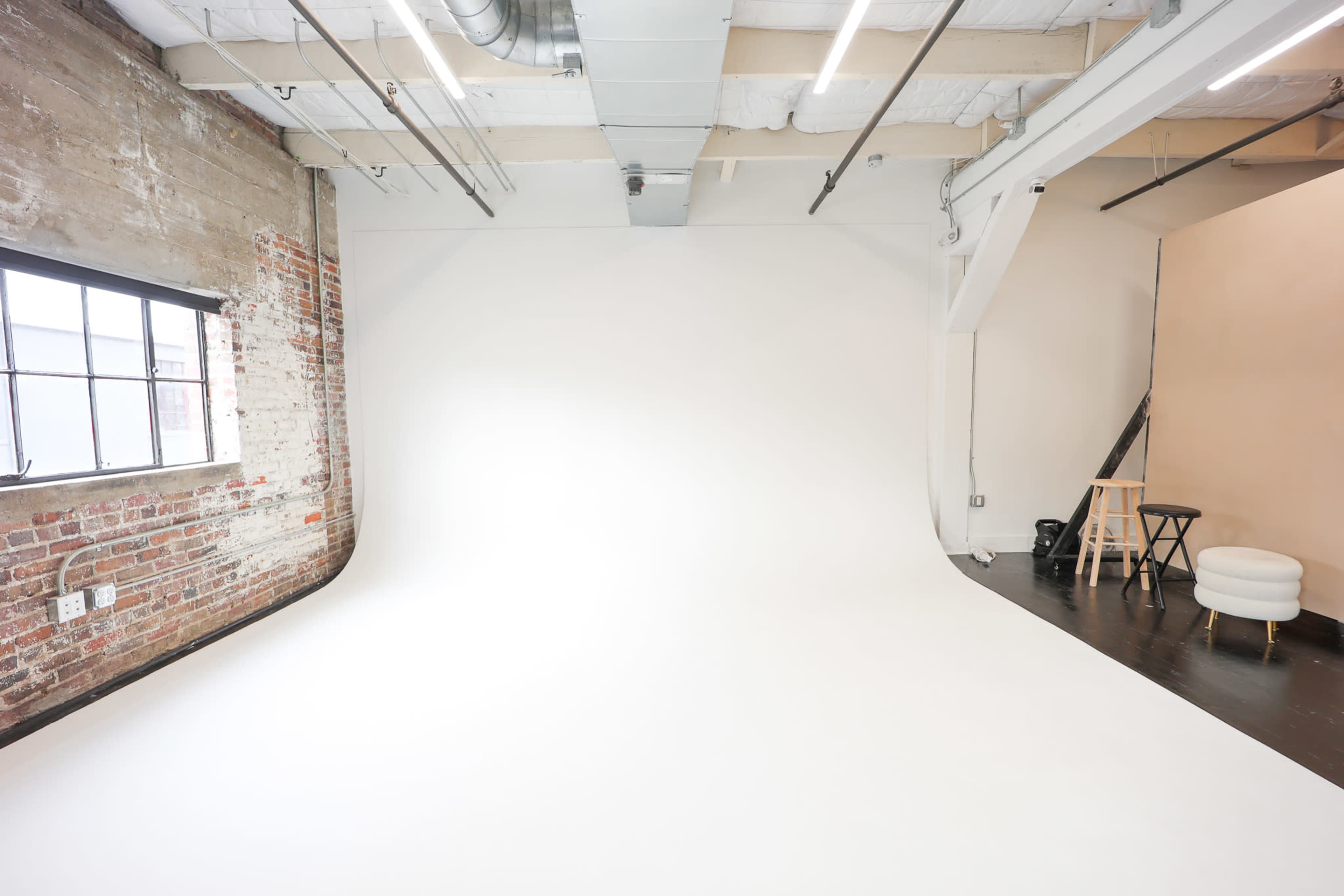 The image shows a spacious photography studio with a white backdrop, exposed brick walls, and a few pieces of furniture in the corner.