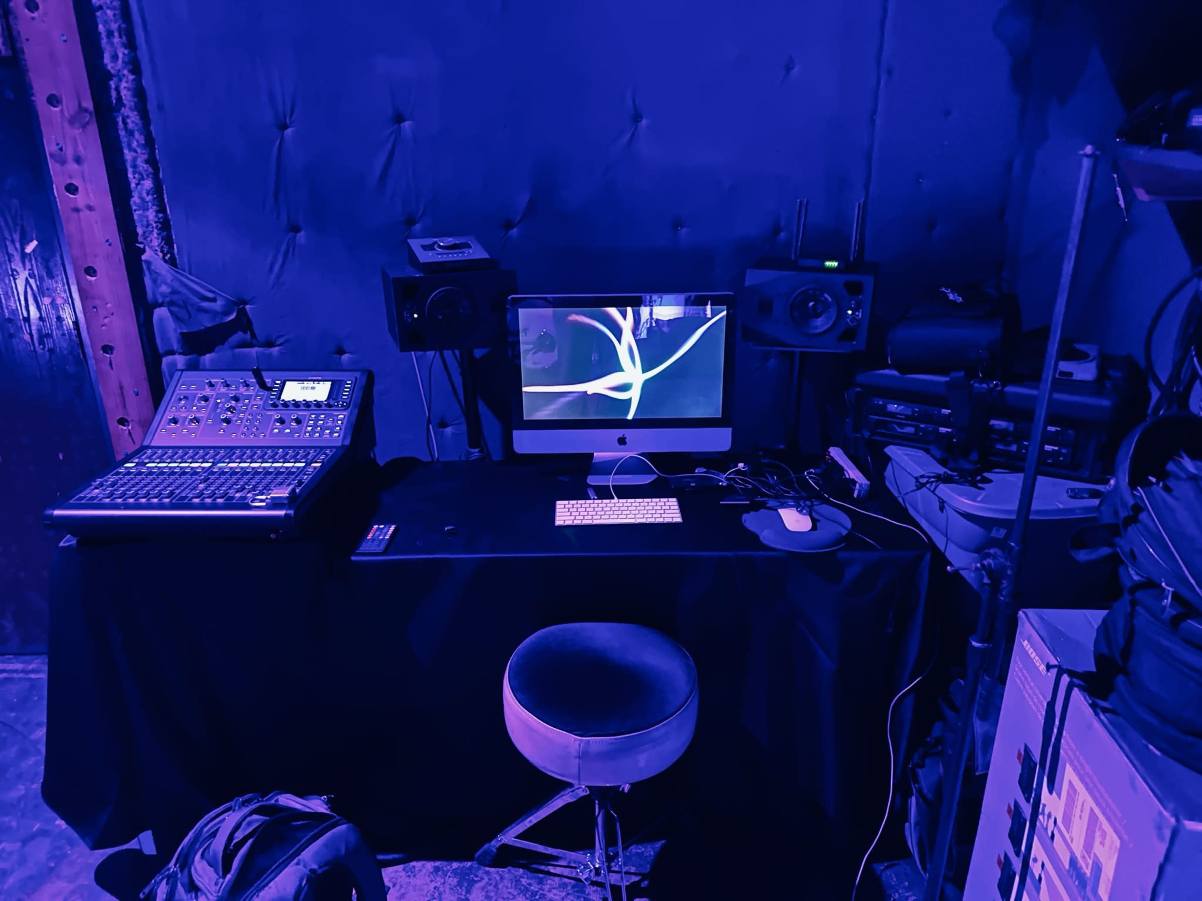 A music production setup featuring a computer, audio interface, speakers, and a stool, all within a dimly lit room.