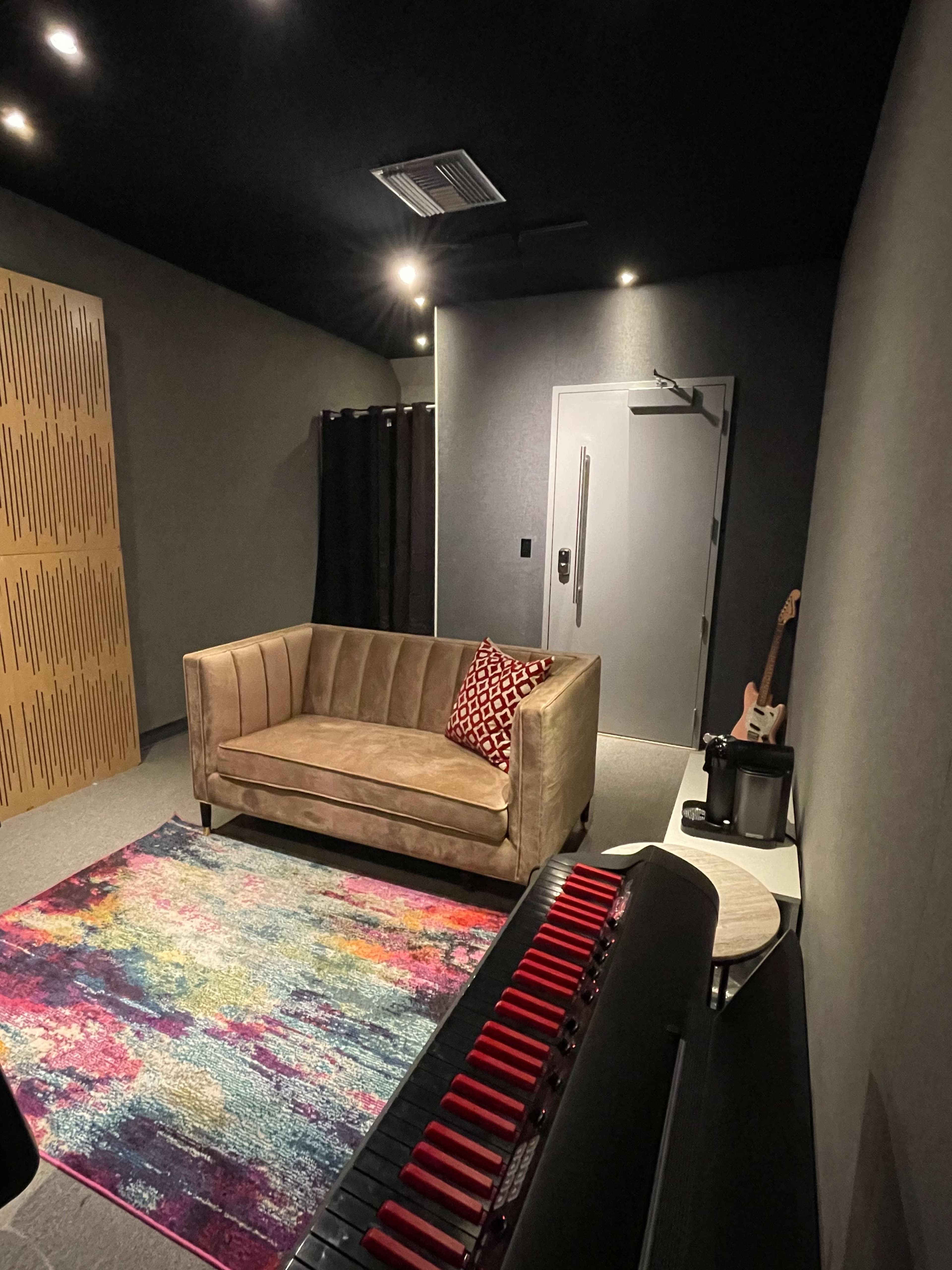 A cozy music studio features a sofa, colorful area rug, and a keyboard in a dimly lit room with soundproof walls.