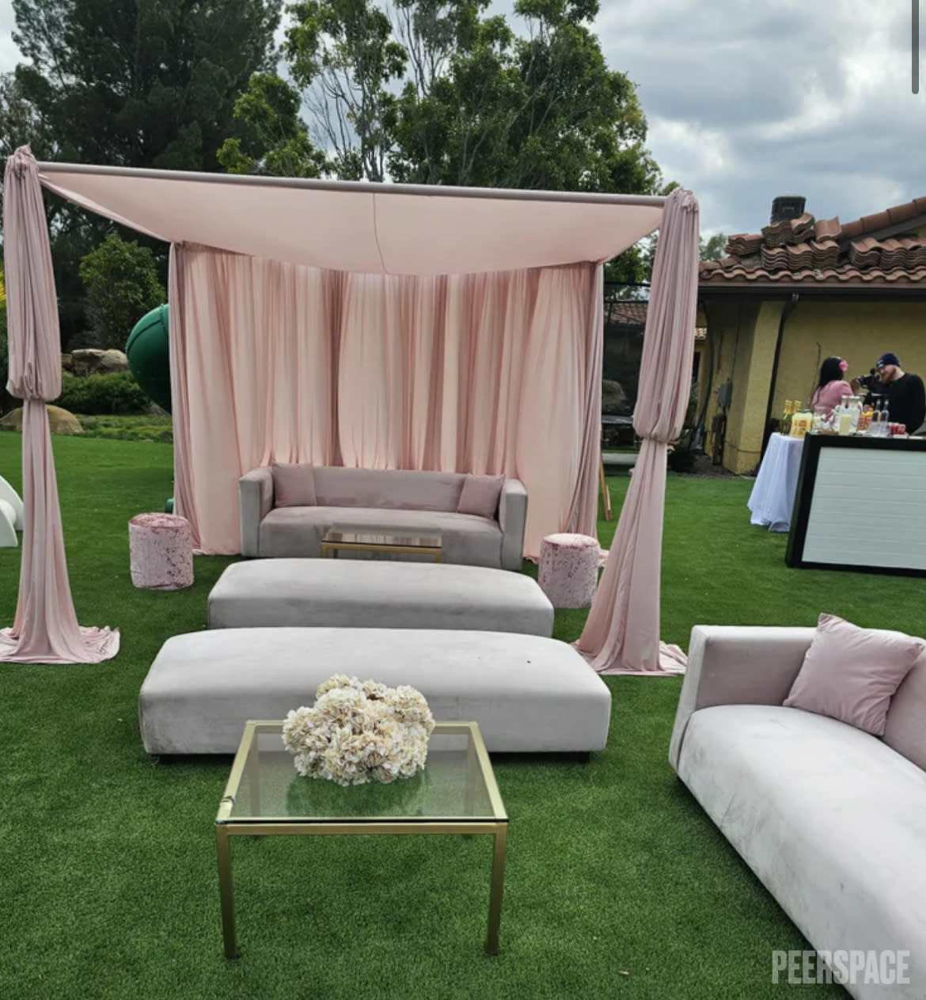 The image shows a stylish outdoor lounge area featuring a light pink draped canopy, soft sofas, and a glass-top coffee table adorned with flowers.