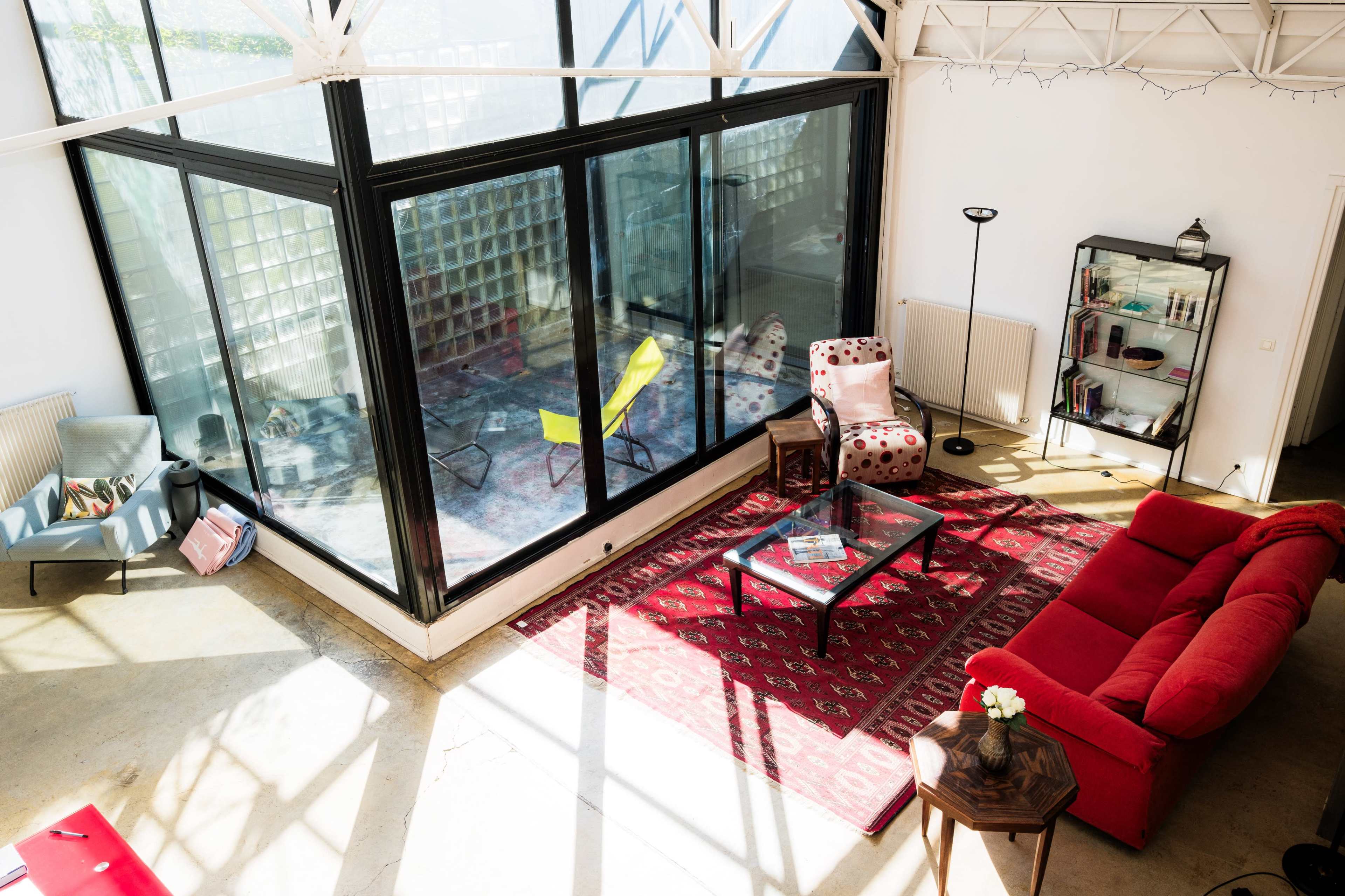 The image shows a modern living space featuring a red sofa, a glass-walled room with colorful chairs, and a patterned rug on a concrete floor.