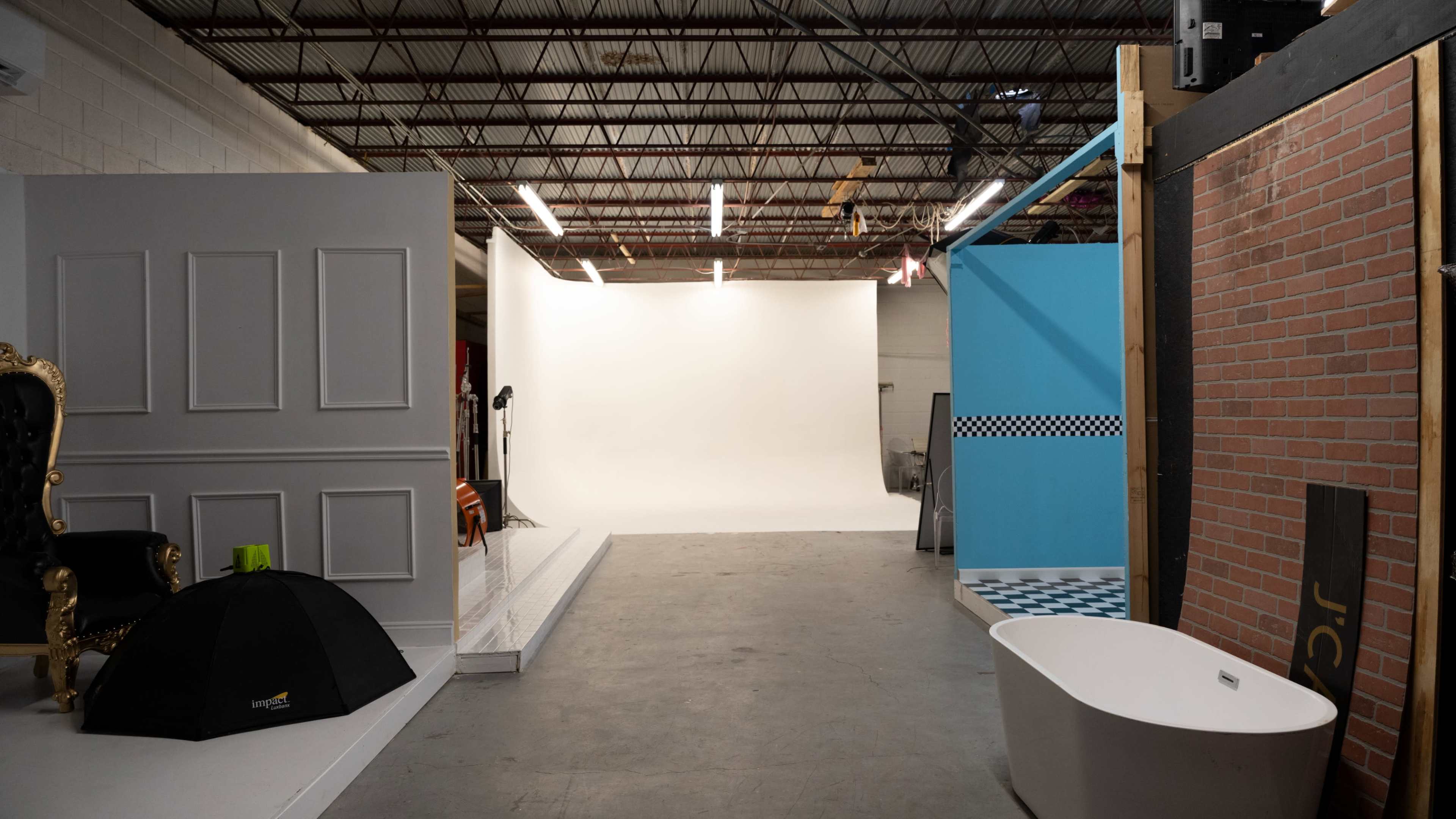 A spacious studio with a blank white backdrop, a black and white checkered floor section, a bathtub, an ornate chair, and various wall sections.