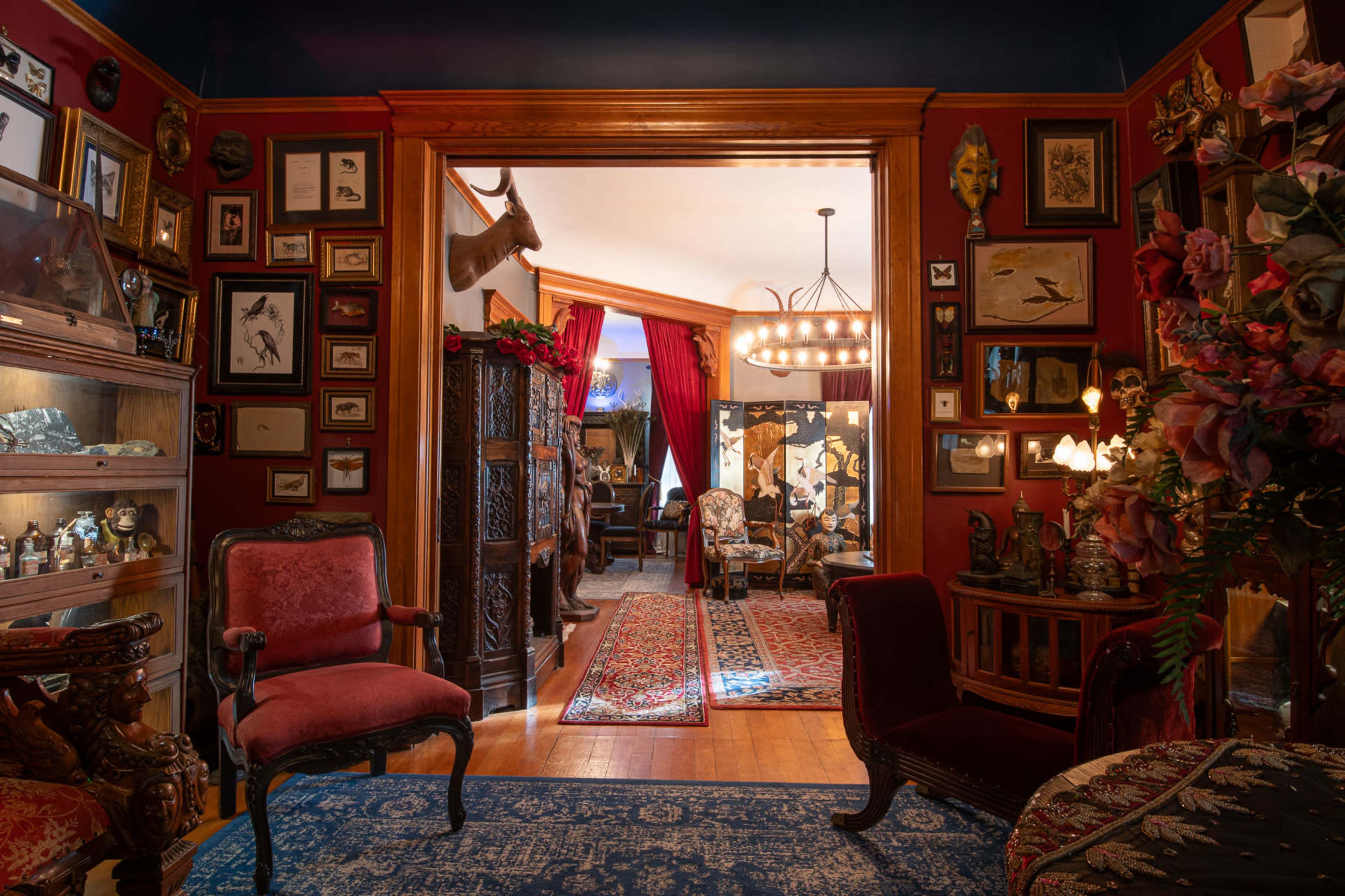 A richly decorated room features ornate wooden furniture, framed wall art, and a view into another elegantly furnished space.
