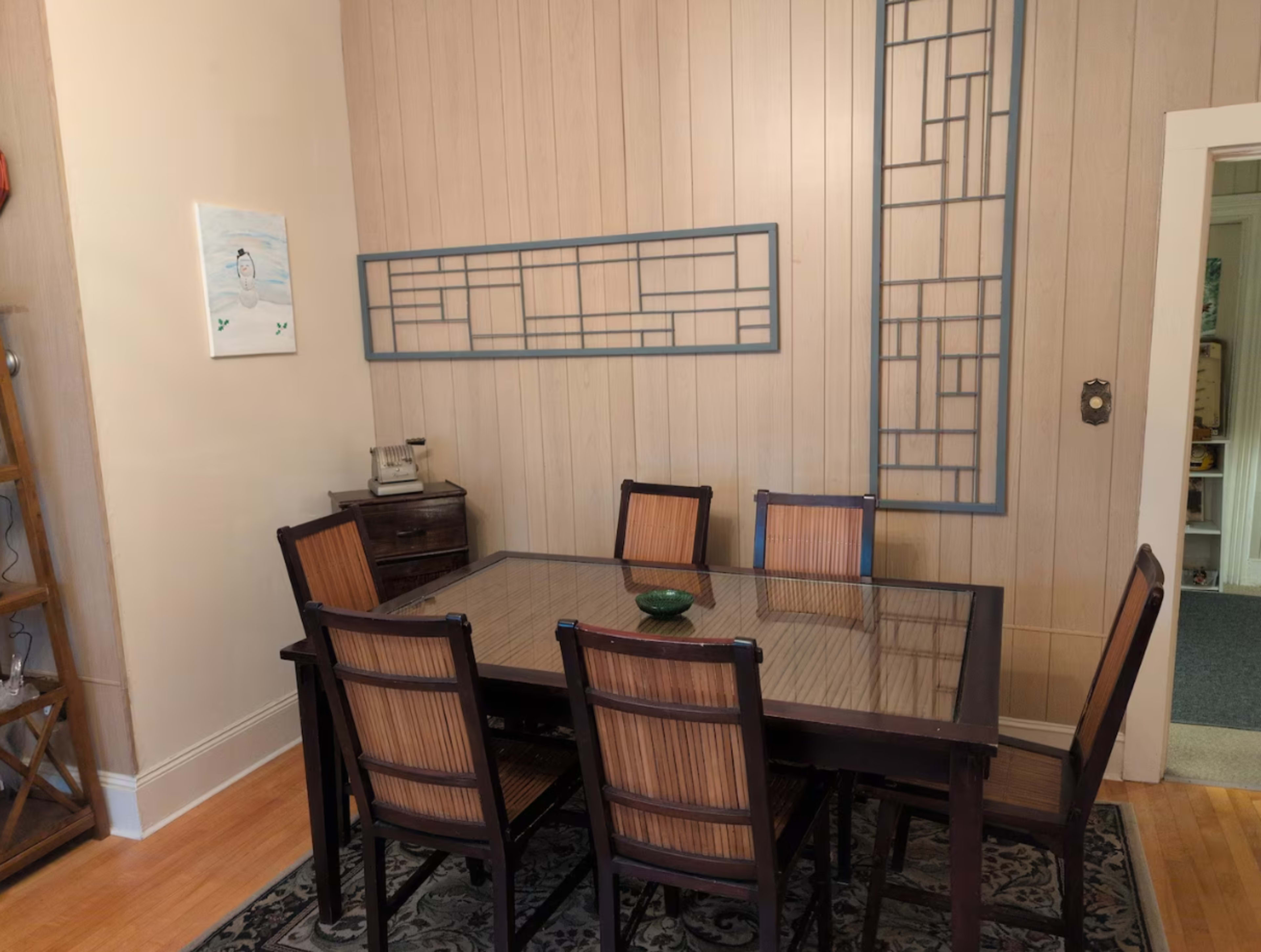 The image shows a dining room with a wooden table surrounded by six chairs, set against a light wood-paneled wall featuring a decorative grid design.