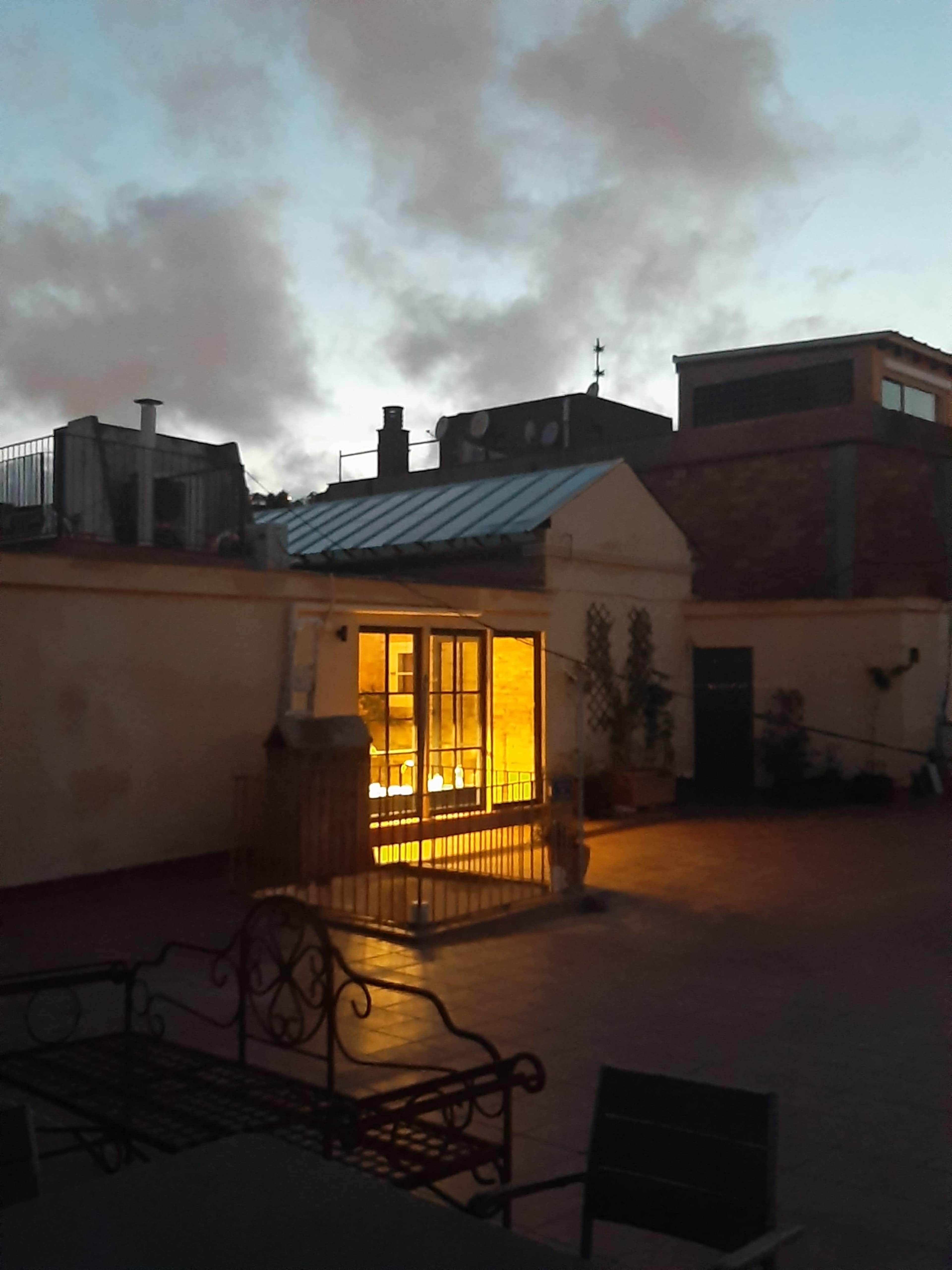 The image shows a rooftop scene at dusk, with a well-lit window in a building and various rooftops in the background.