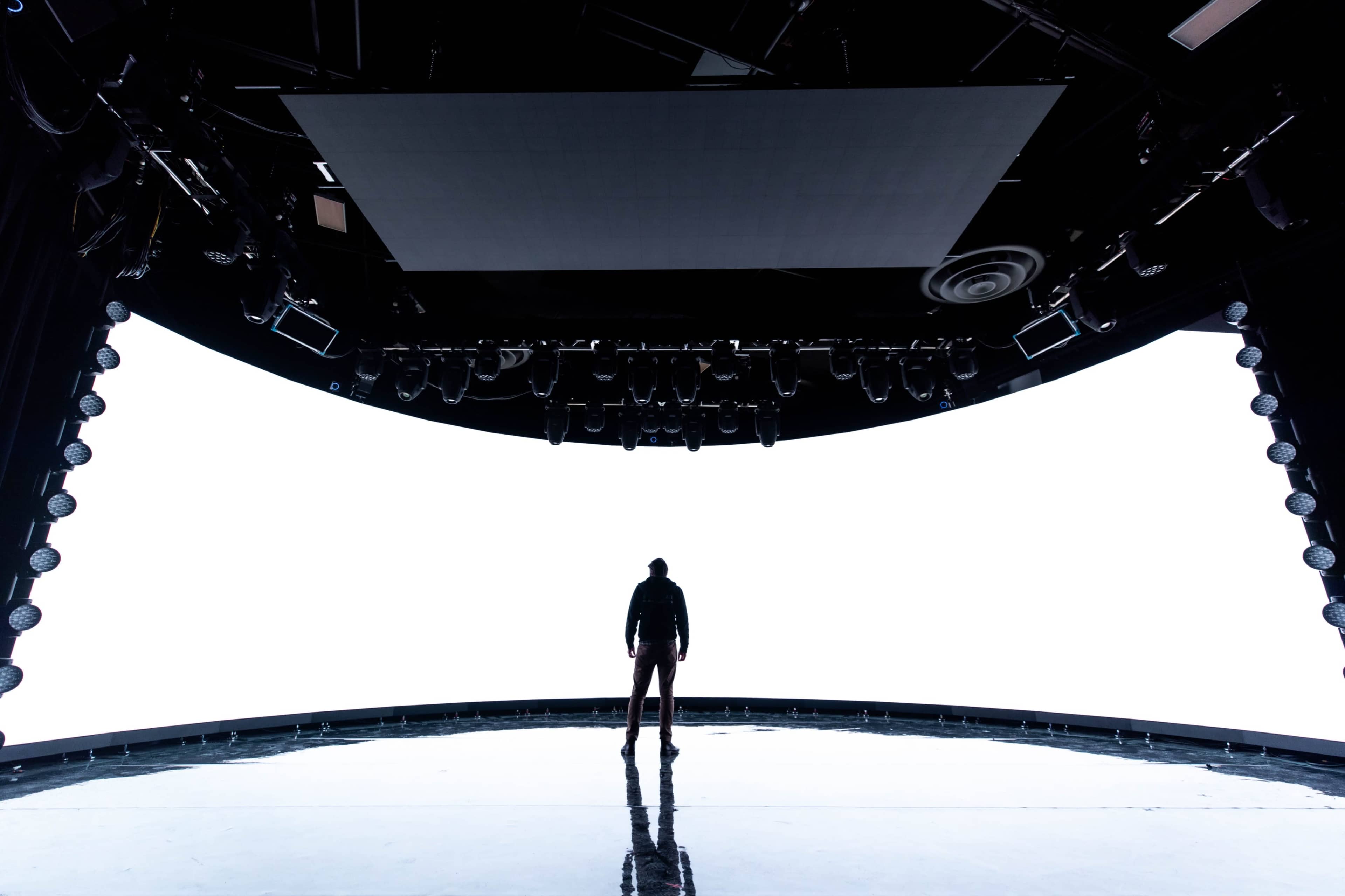 A person stands alone on a dimly lit stage with a large blank screen behind them.